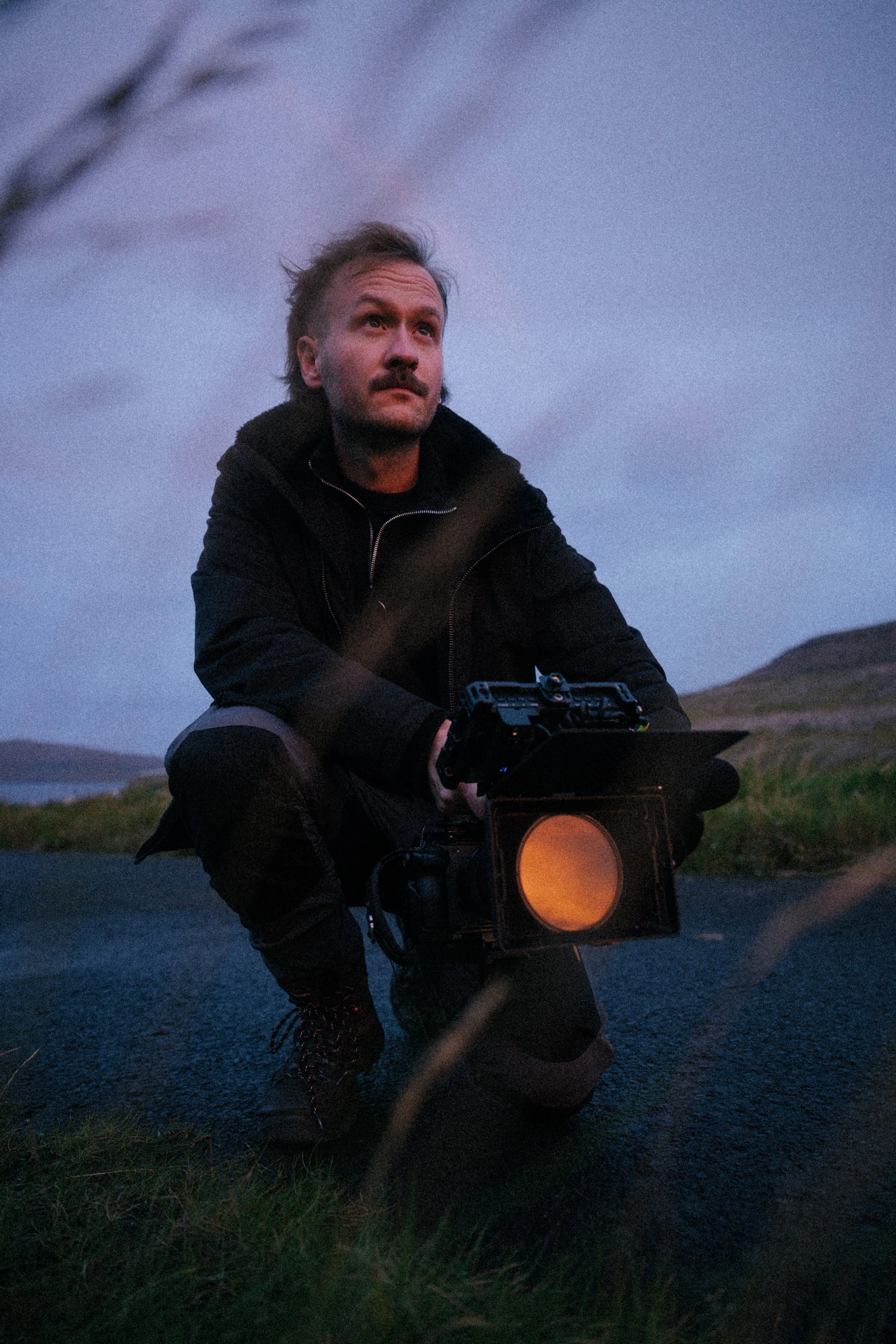 A man kneeling outdoors at dusk, holding a camera with a large filter, with rolling hills and a cloudy sky in the background.