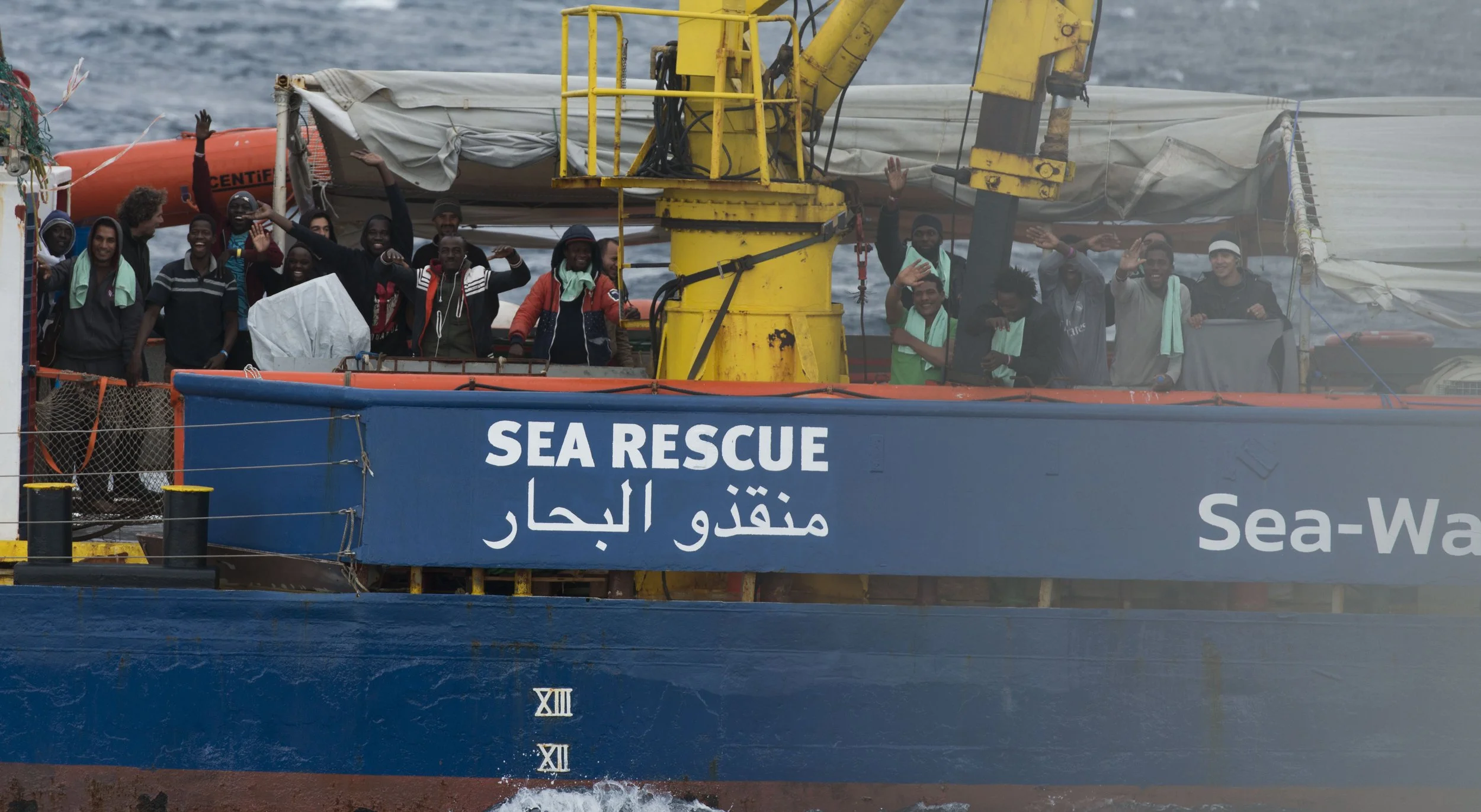 Group of people onboard a sea rescue boat waving and smiling. The boat has a blue hull with 'SEA RESCUE' written in English and Arabic. The water is visible in the background.
