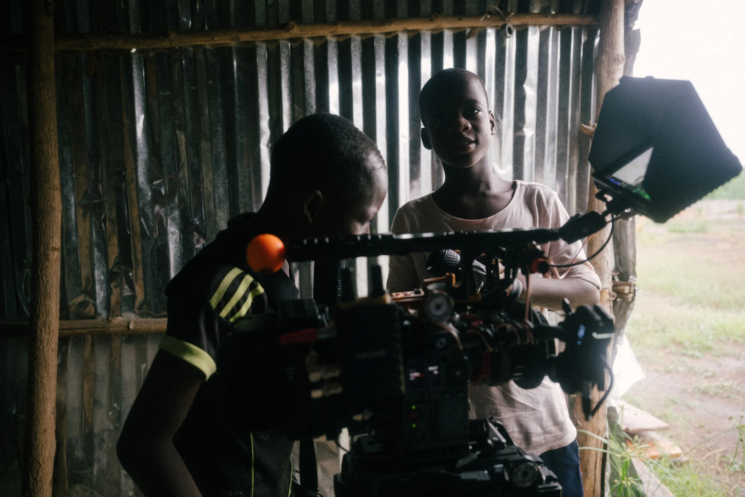 Two children, a boy and a girl, are filming with a large professional video camera inside a rustic wooden and metal shed, with natural light coming from outside.