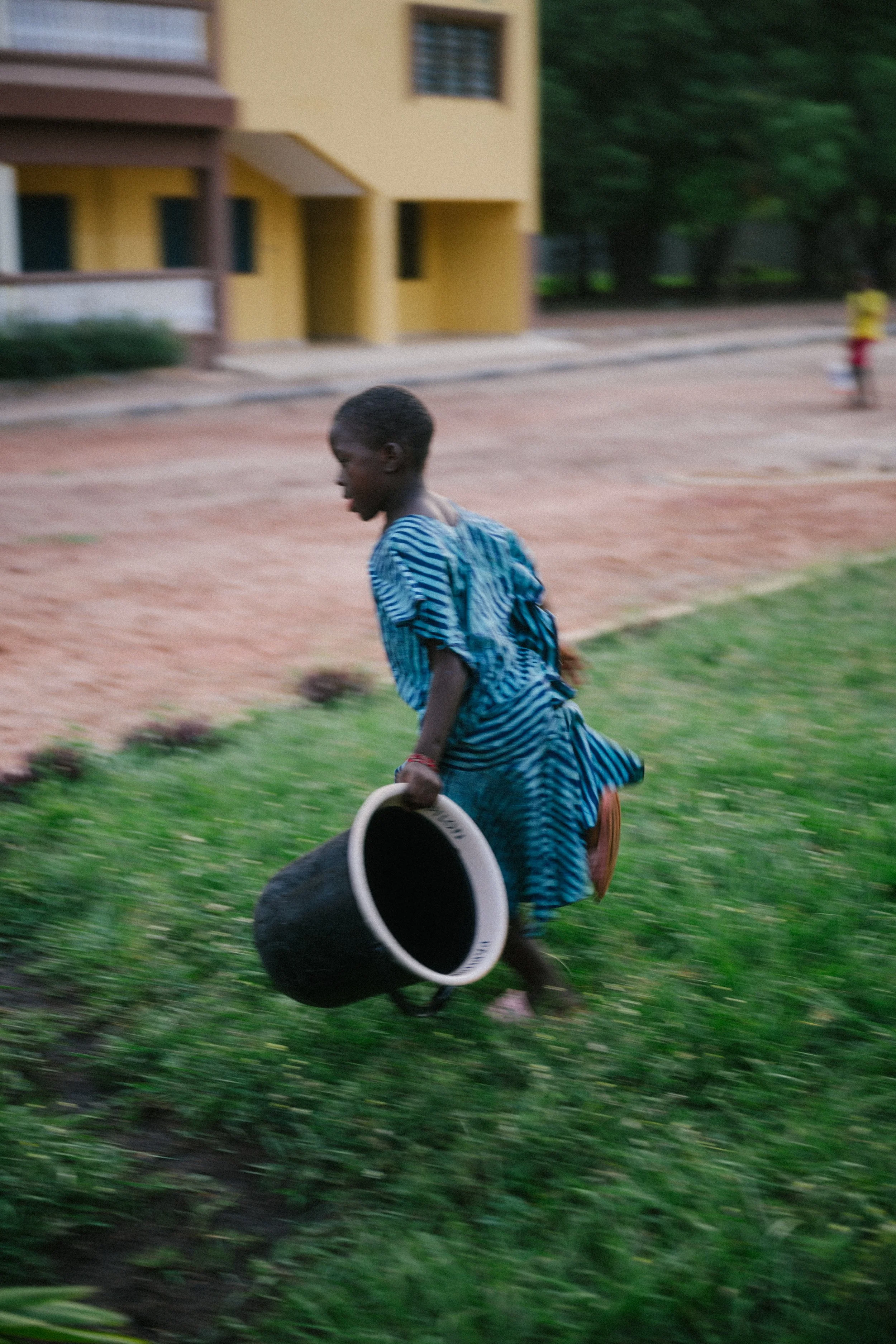 A young boy in a striped blue dress running outdoors while holding a black bucket.