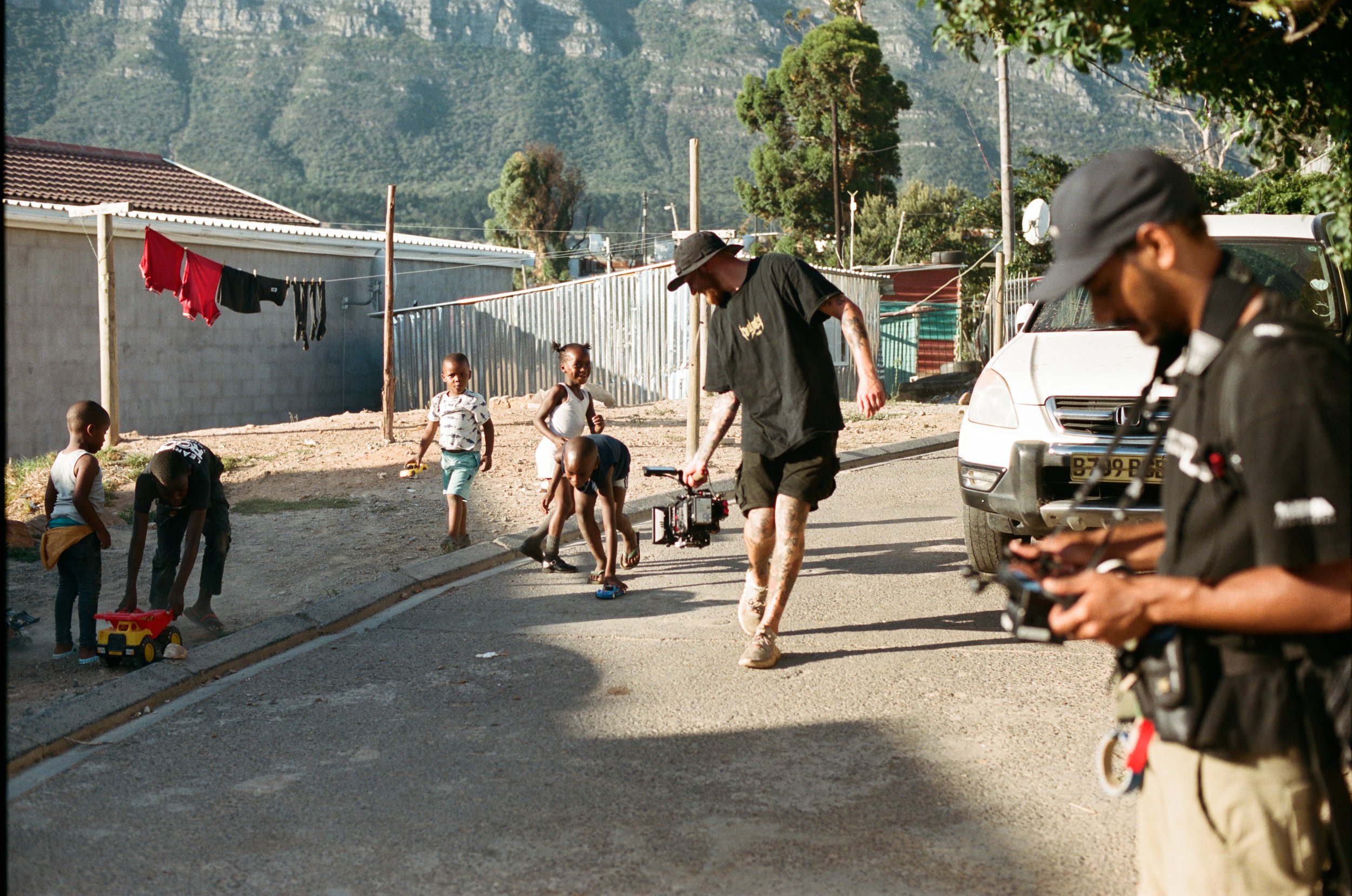 Behind-the-scenes scene showing a film crew filming children playing outside on a quiet street, with mountains and a mix of houses and fences in the background.