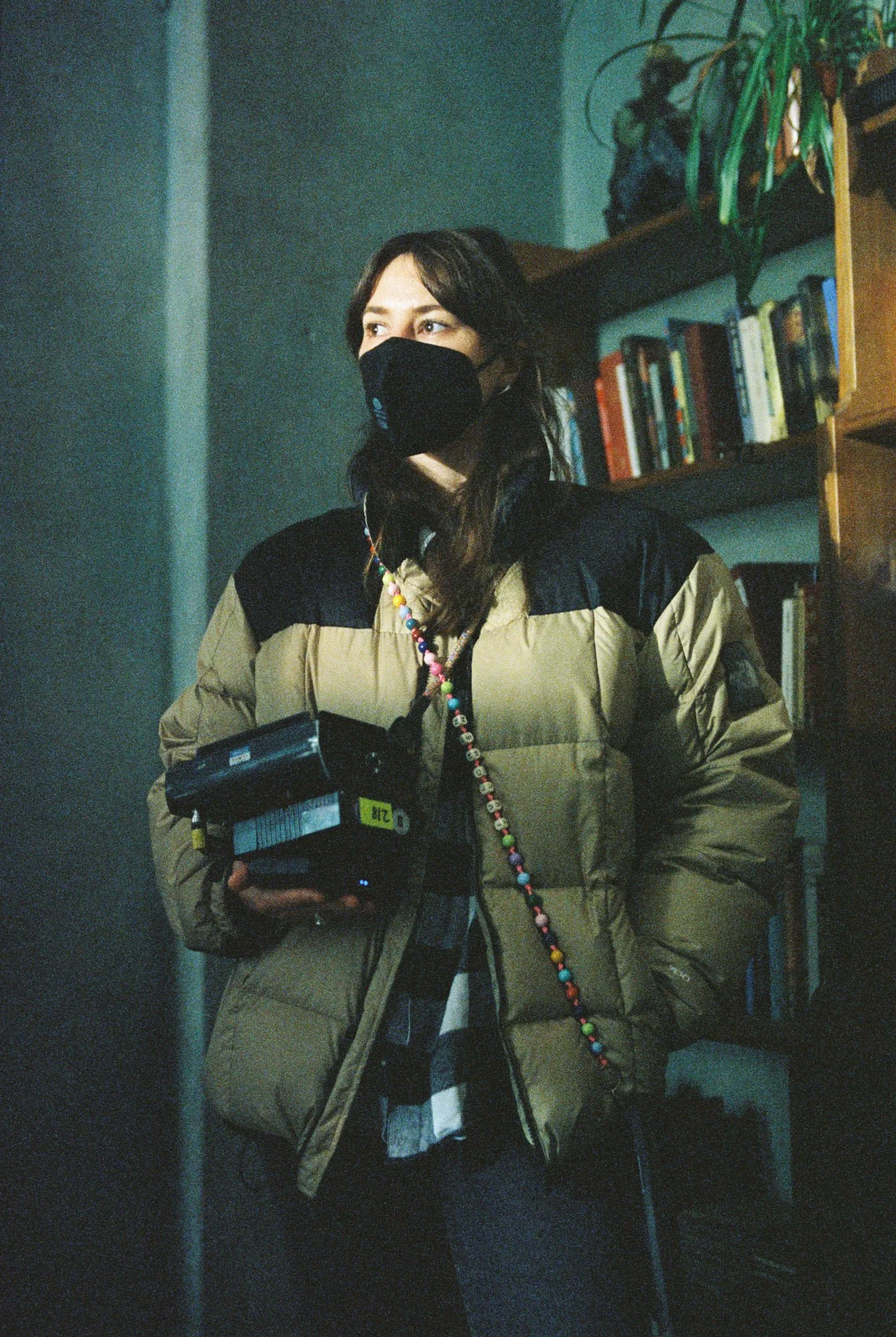 A woman wearing a black face mask, a tan and black puffer jacket, holding a camera, with a colorful beaded strap, standing in a room with bookshelves and a plant.