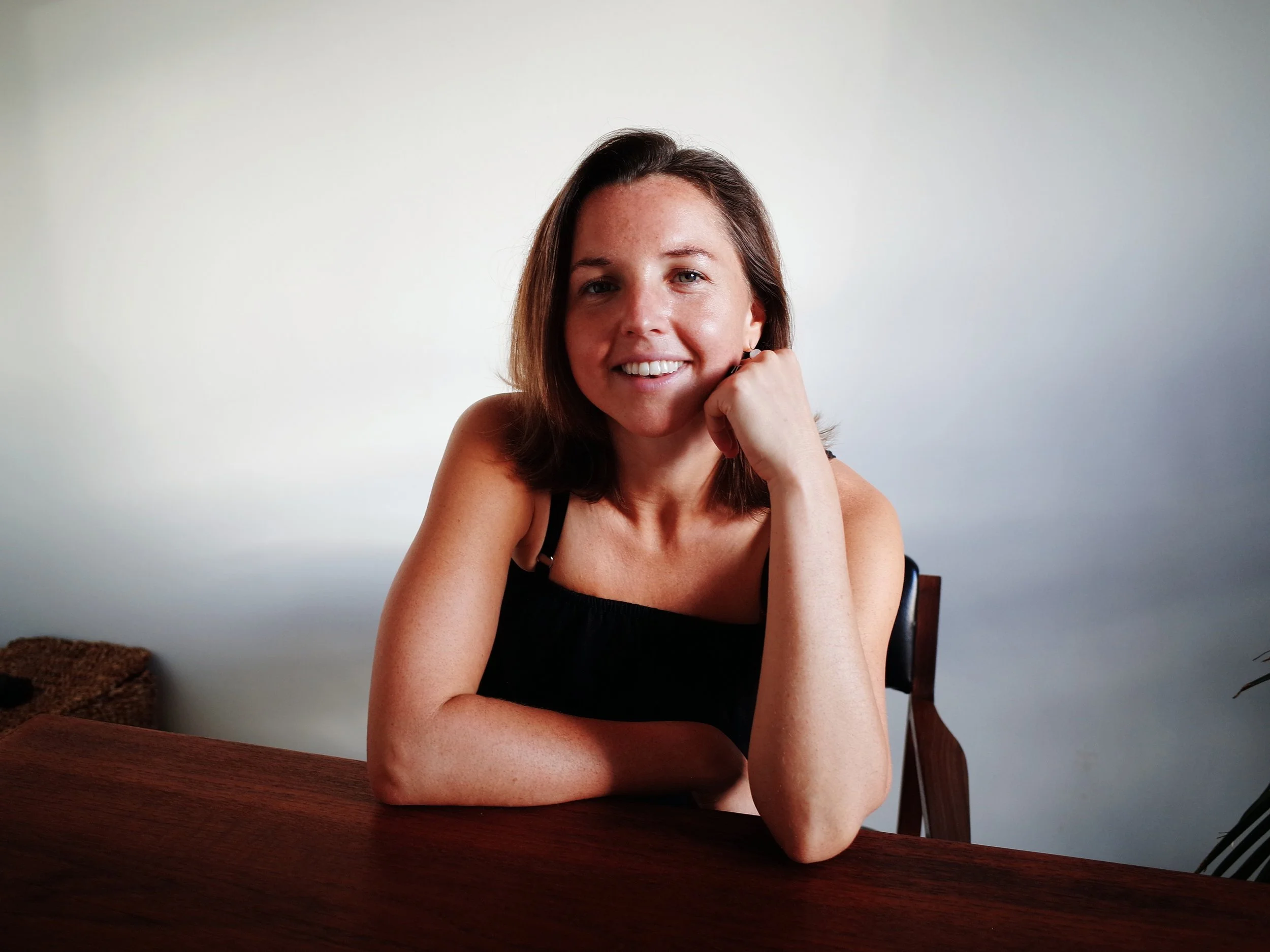 A smiling woman with shoulder-length brown hair, sitting at a wooden table with a casual black sleeveless top, resting her chin on her right hand, in a minimalistic setting with neutral-colored walls.