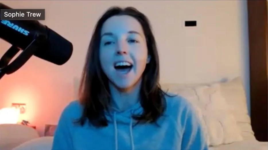 A young woman with brown hair, wearing a gray hoodie, is speaking into a microphone during a video call or recording. She is smiling and appears to be happy.