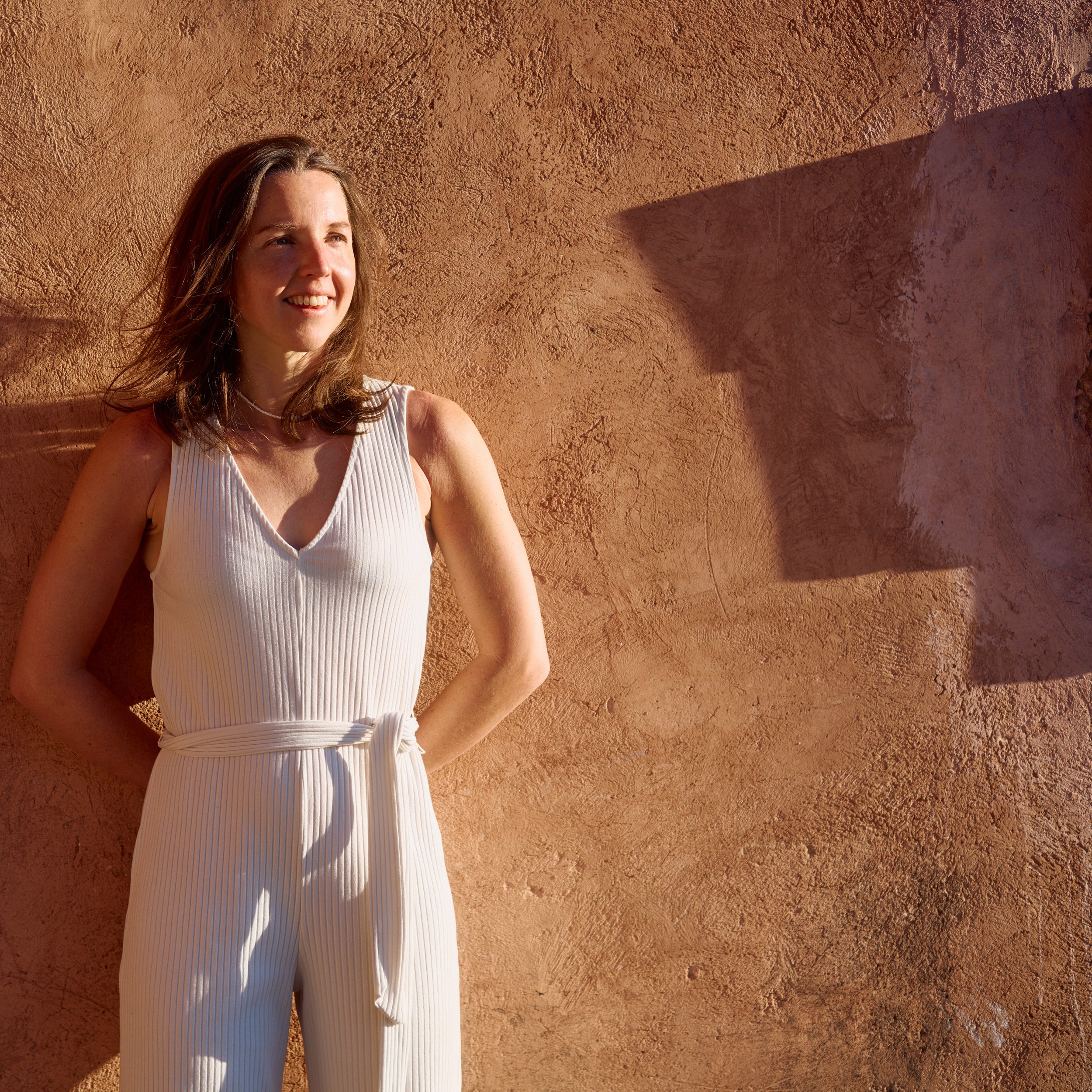 A woman with long brown hair, smiling, wearing a sleeveless white dress with vertical pleats, standing against a textured terracotta wall with a shadow cast on it.
