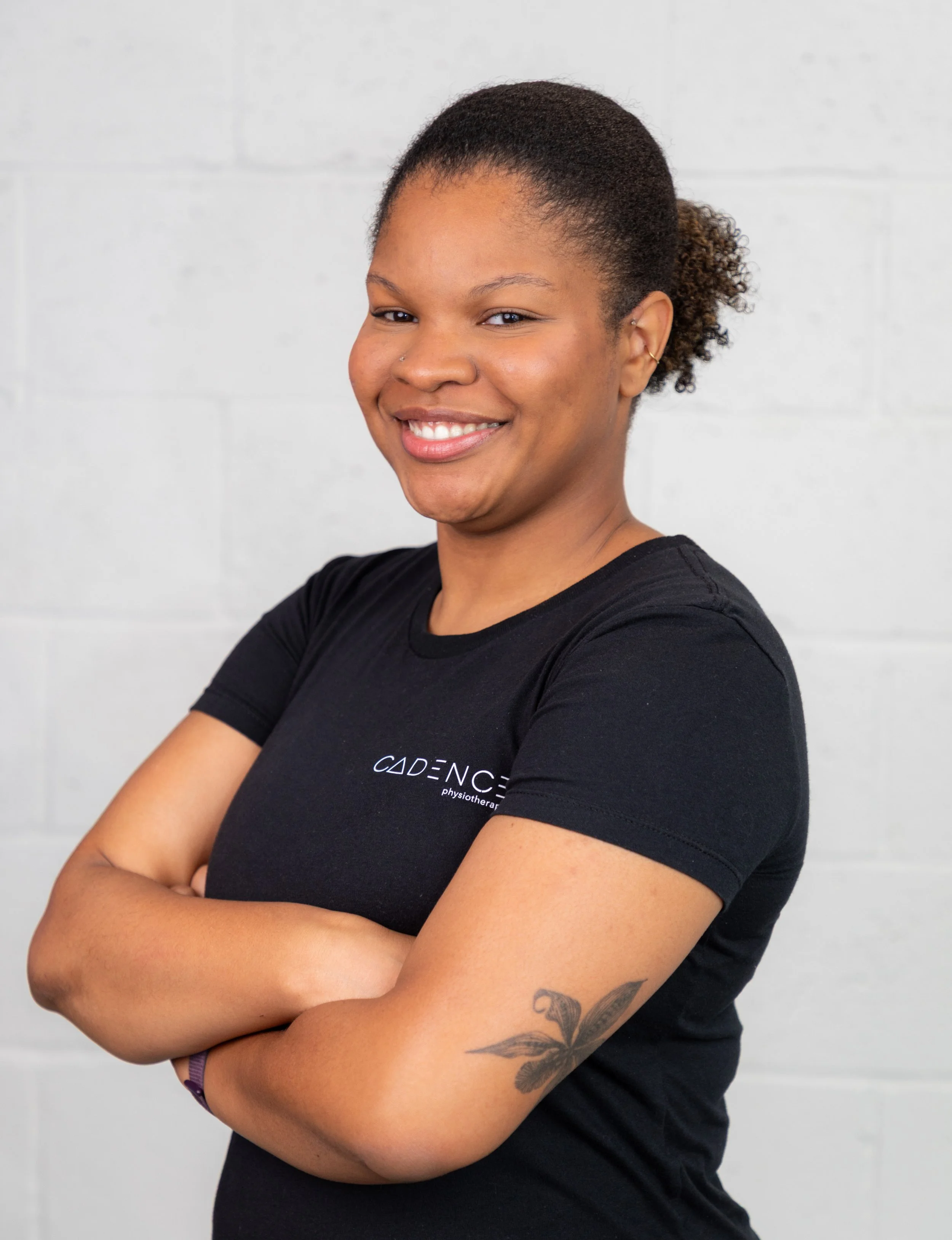 A young woman with dark hair tied back, smiling, standing against a white brick wall, wearing a black t-shirt with 'CADENCE' and 'physiotherapy' written on it, arms crossed.