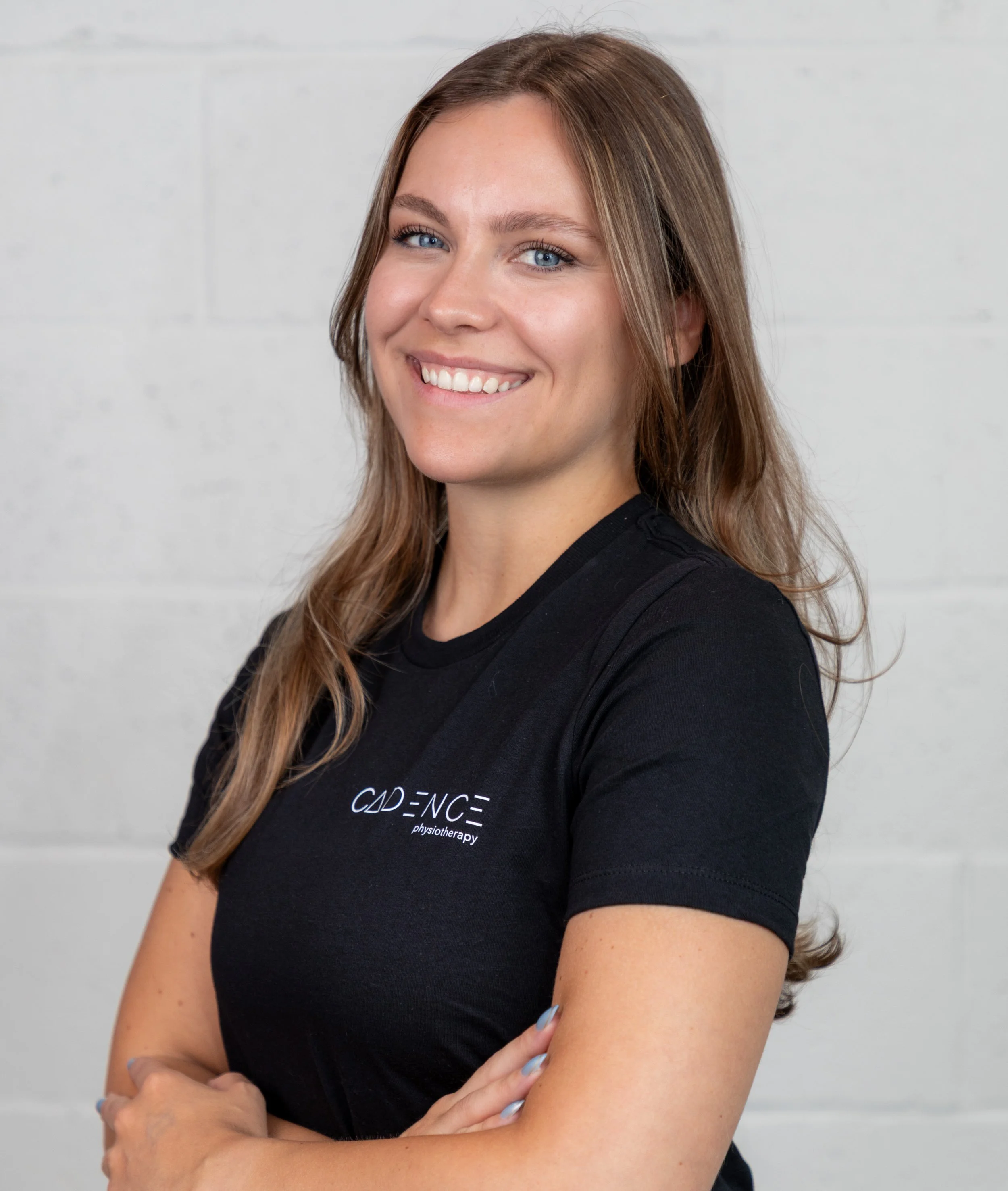 A smiling woman with long blonde hair standing against a white brick wall, wearing a black t-shirt with the logo 'CADENCE physiotherapy'.