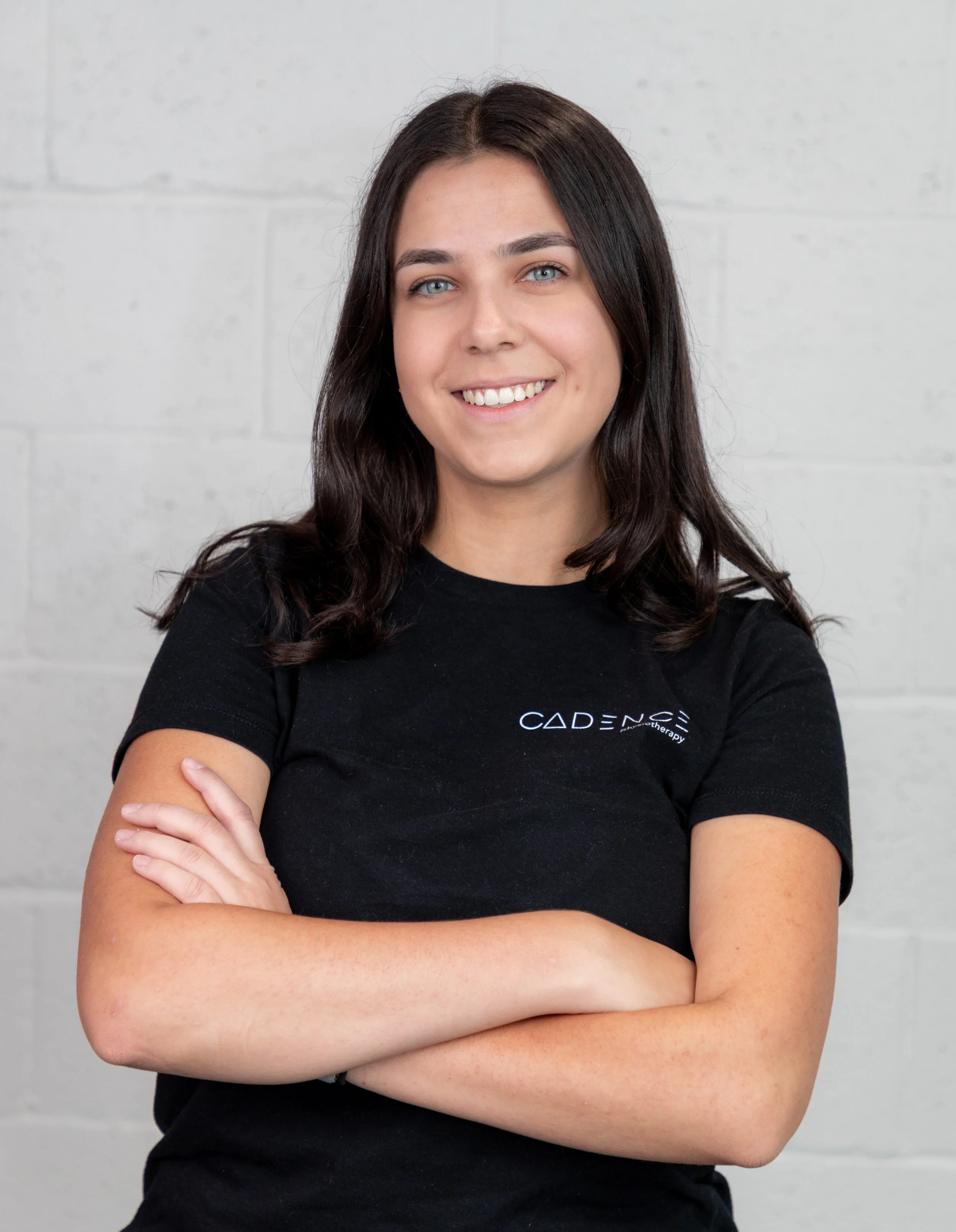 A young woman with long dark hair, wearing a black t-shirt, standing against a plain white wall, smiling softly.