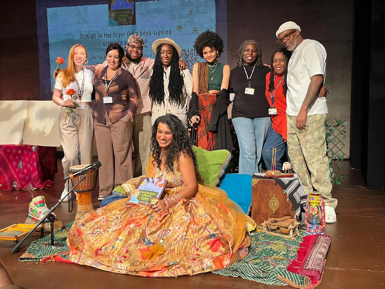 Author Brwn Girl in the Ring pictured holding her book on the stage of the Belgrade Theatre with her creative collaborators Emily, Amy, Reisz, Elizabeth, Malikah, Steady, and BSL interpreters Jacqueline & Hyacinth standing behind her.