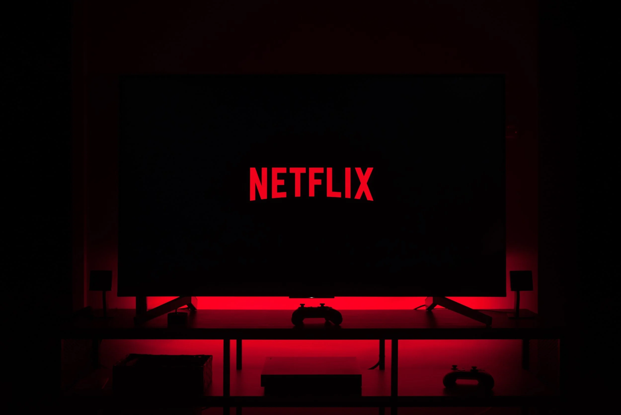 A television screen displaying the Netflix logo with red illumination around it and a gaming controller on a table in front.