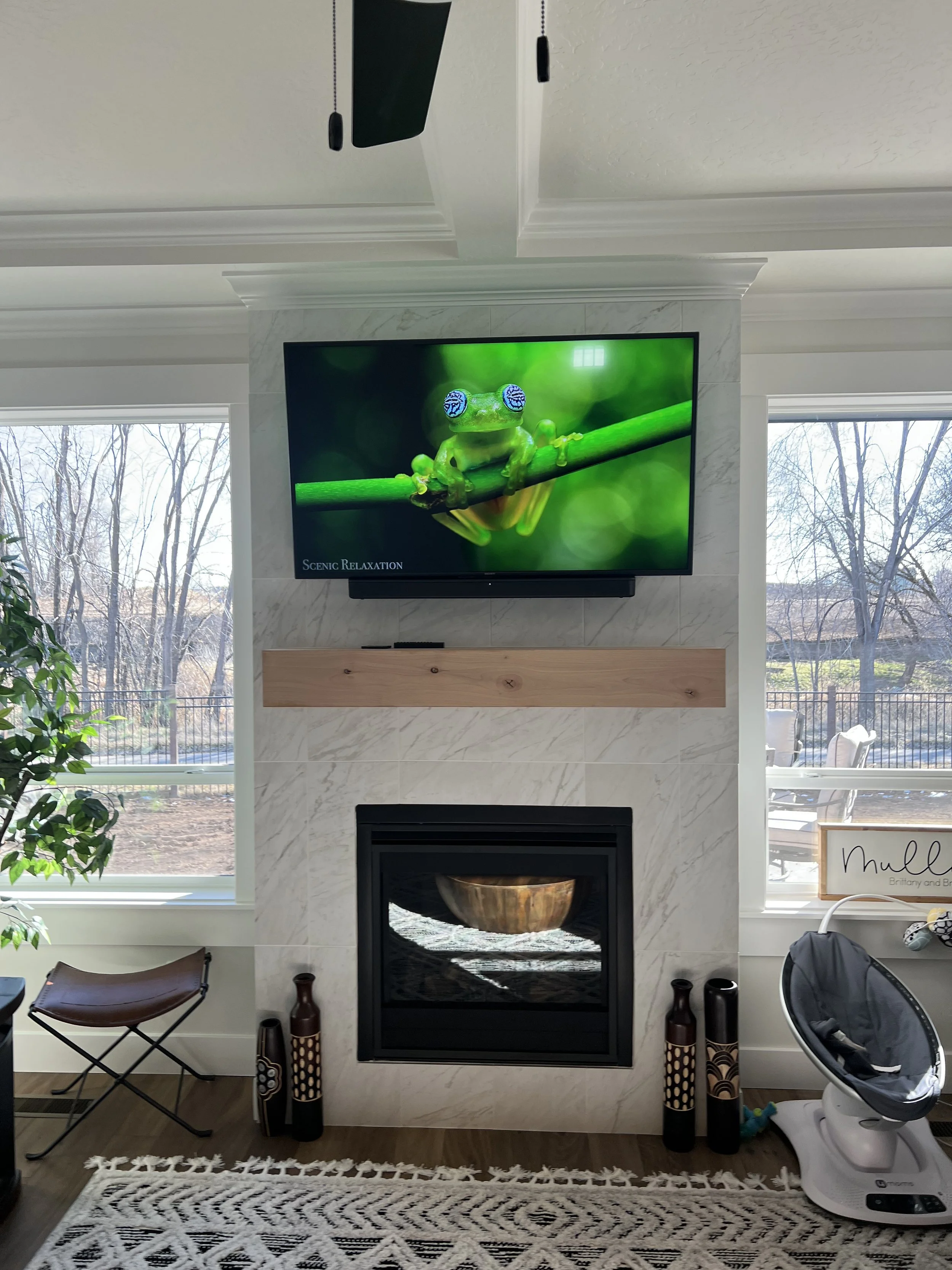 Living room with a mountain-style fireplace, a mounted flat-screen TV showing a frog, and large windows showing outdoor trees and a pool.