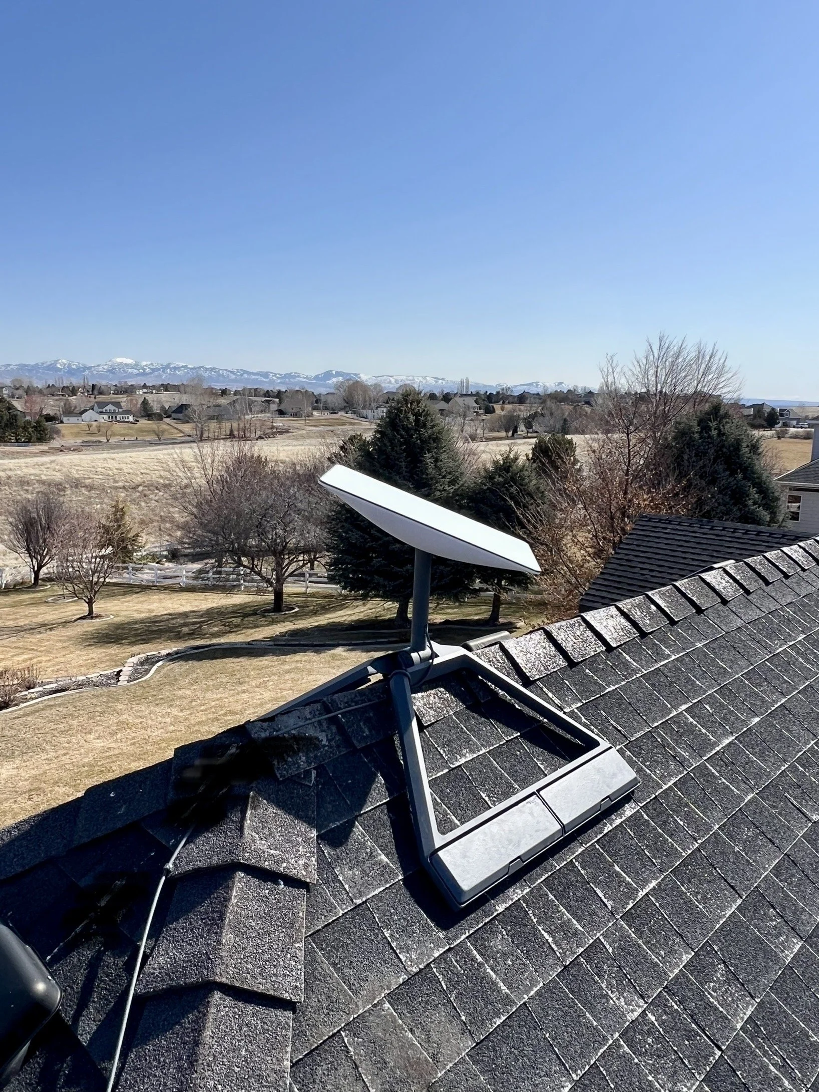A rooftop with satellite dish and cable wire, overlooking a backyard with trees and open fields, with mountains in the background under a clear blue sky.