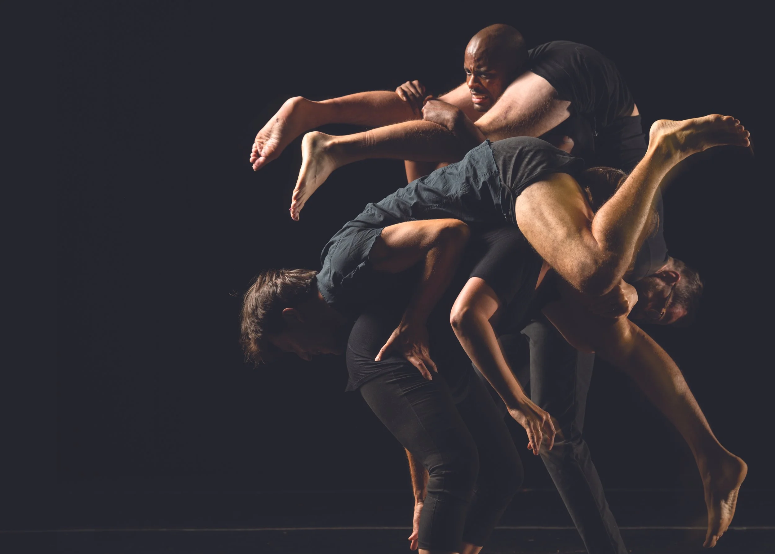 Against a black background, two men dressed in black carry their partners over their shoulders