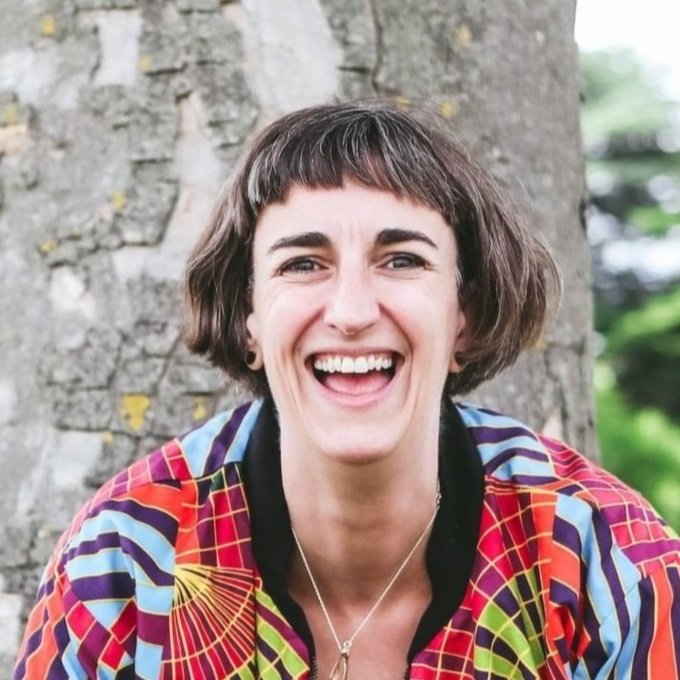 A woman with short, dark hair and a big smile, wearing a colorful patterned jacket, standing outdoors in front of a tree.