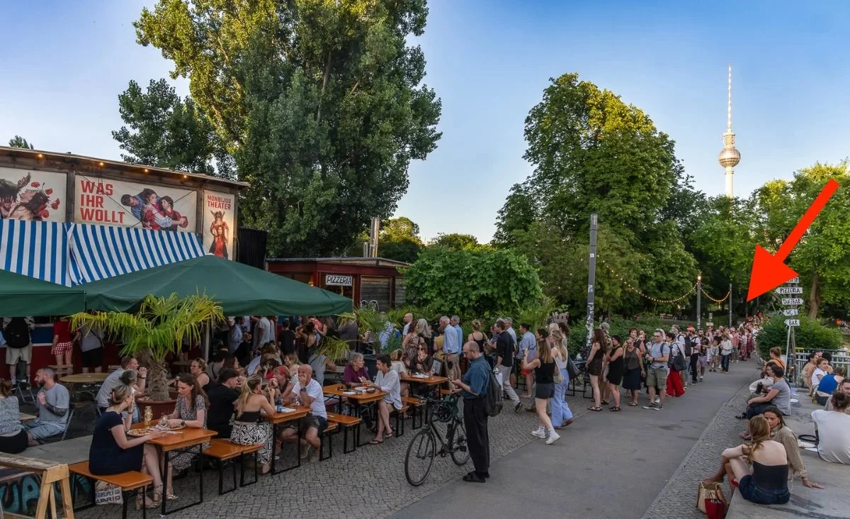 A wide daytime view along Am Zwirngraben with beer garden benches and a striped food stall on the left, a crowd down the path, and a red arrow pointing left