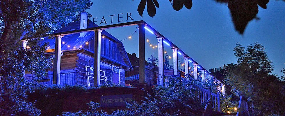 The illuminated timber Wilhelmshütte theater building at twilight, strung with fairy lights, seen from the path leading up to the entrance in Berlin