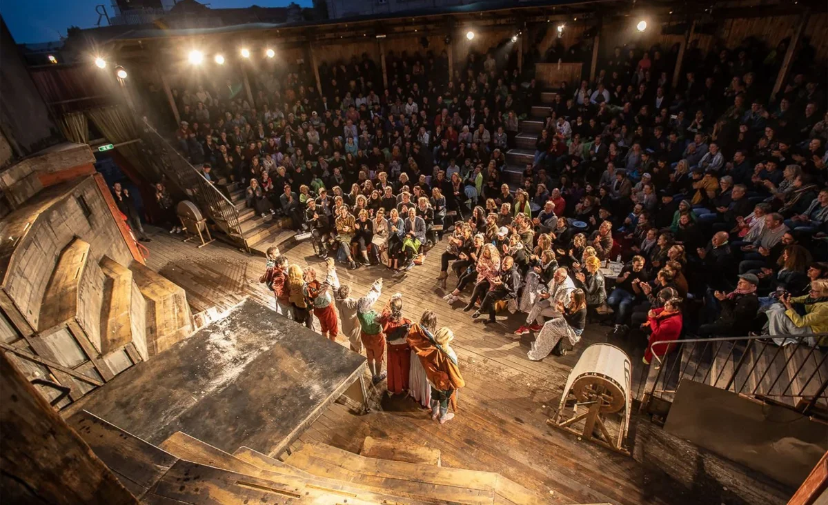 Elevated view of a packed audience surrounding costumed performers on the wooden open-air stage at Monbijou Theater in Berlin