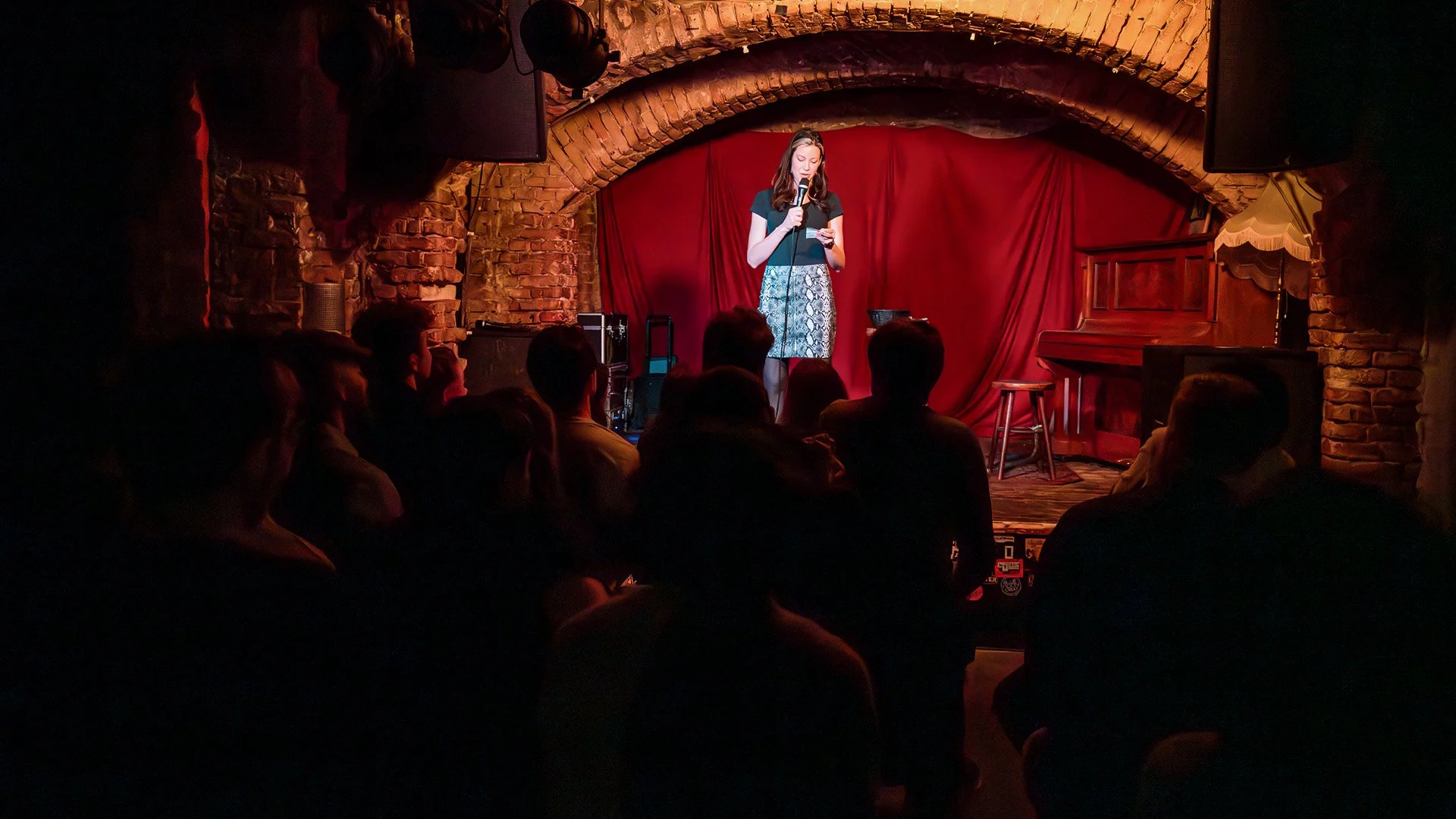 A comedian on a red-curtained stage under a brick arch, seen from behind a close-packed audience in Berlin