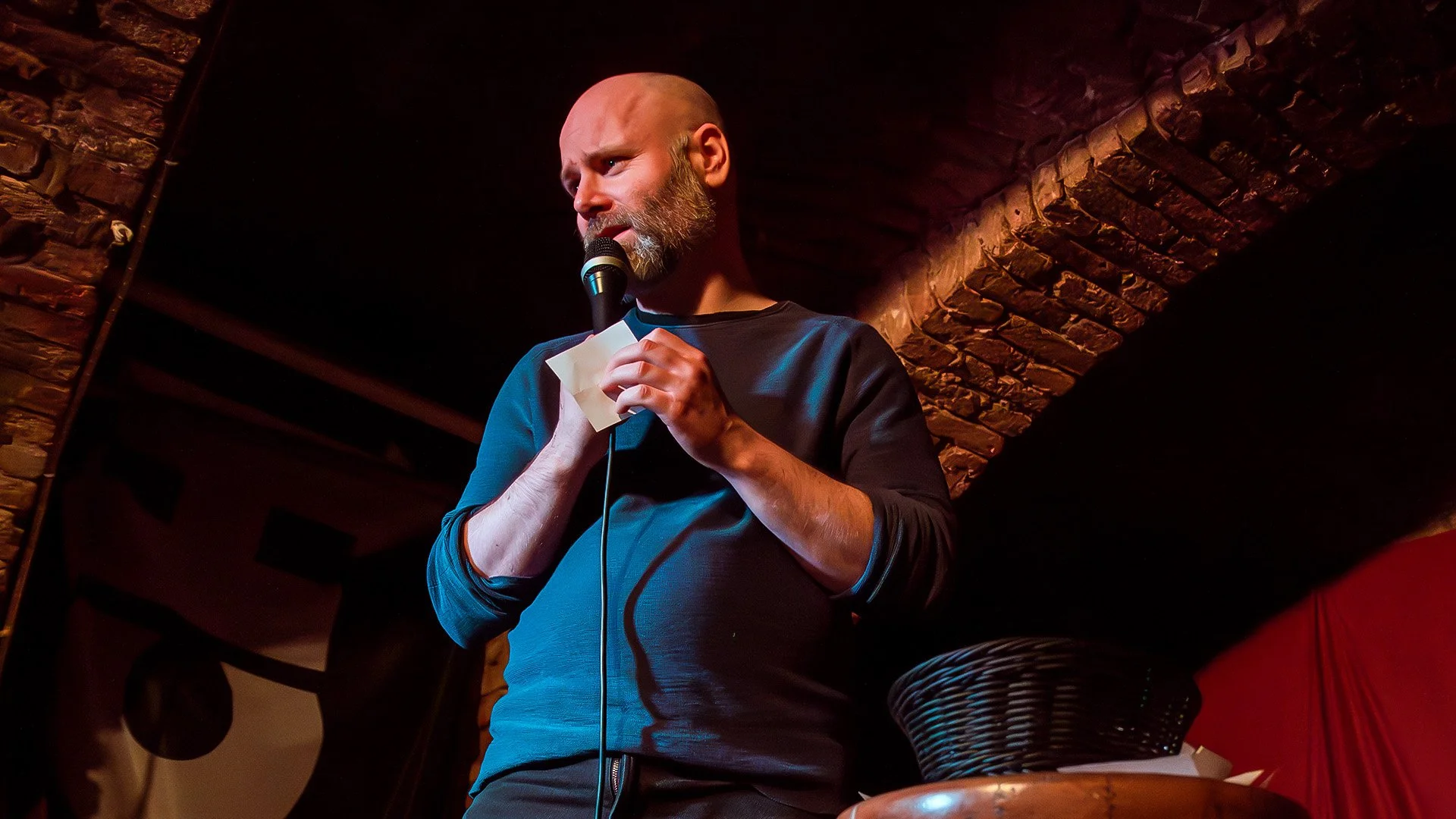 Brendan Hickey reads a note from the audience on stage under a brick arch at an English comedy show in Berlin