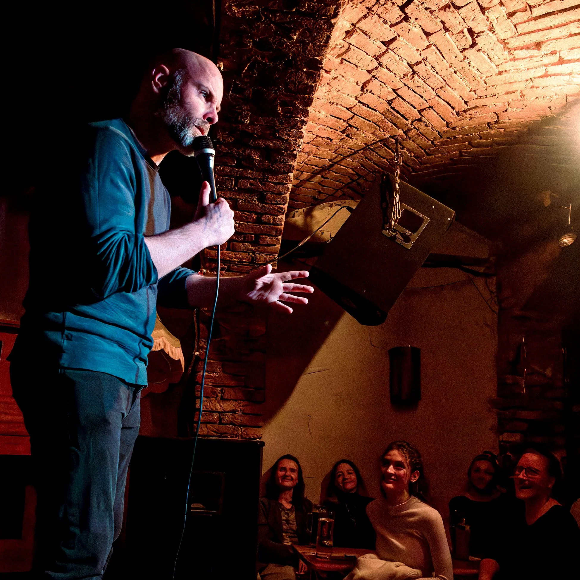 Brendan Hickey (comedian) performing stand-up to a laughing audience in a dark-light brick-vaulted Berlin comedy venue