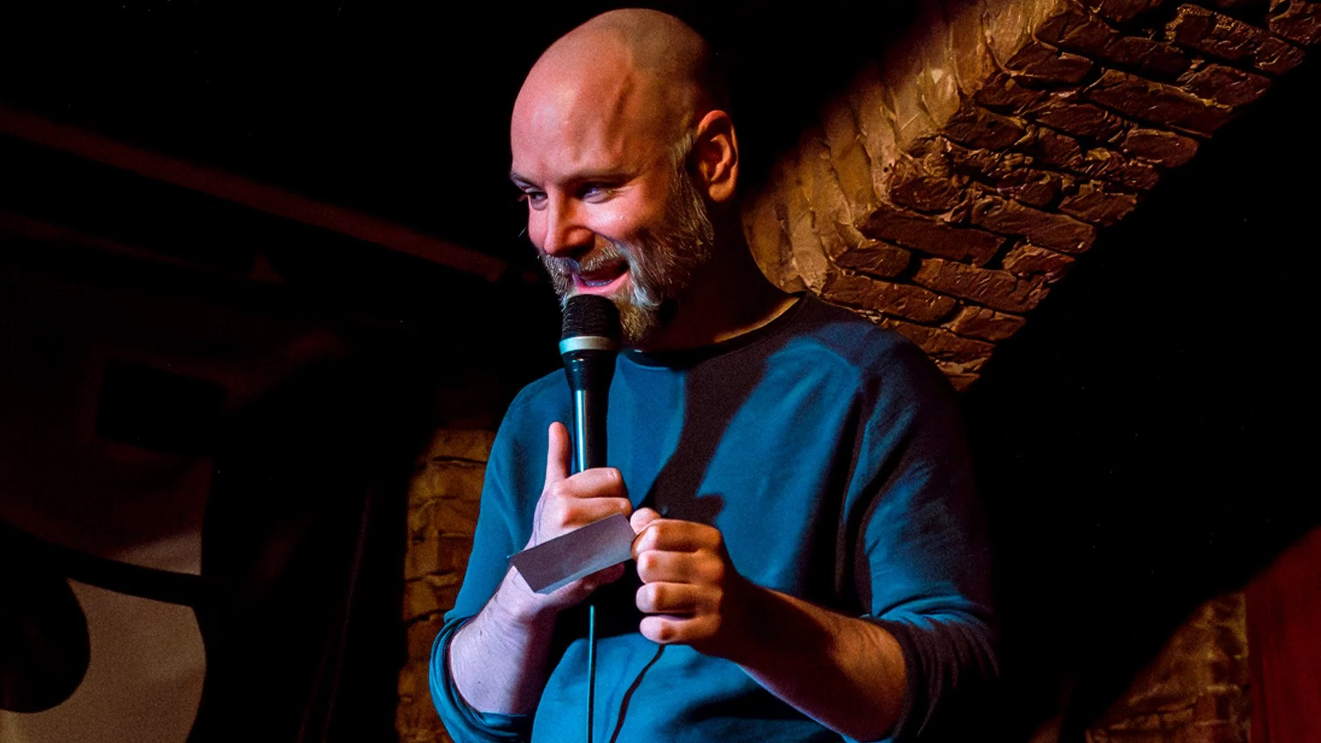 Host Brendan Hickey reads an audience note under a spotlit brick arch at a Darkest Thoughts comedy show in Berlin