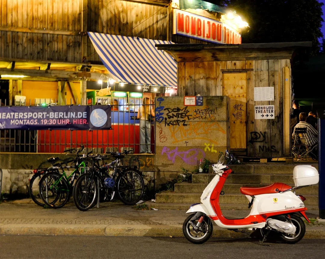 The lit Monbijou Theater sign above the wooden street-front entrance at night, with bikes and a scooter parked outside, in Berlin