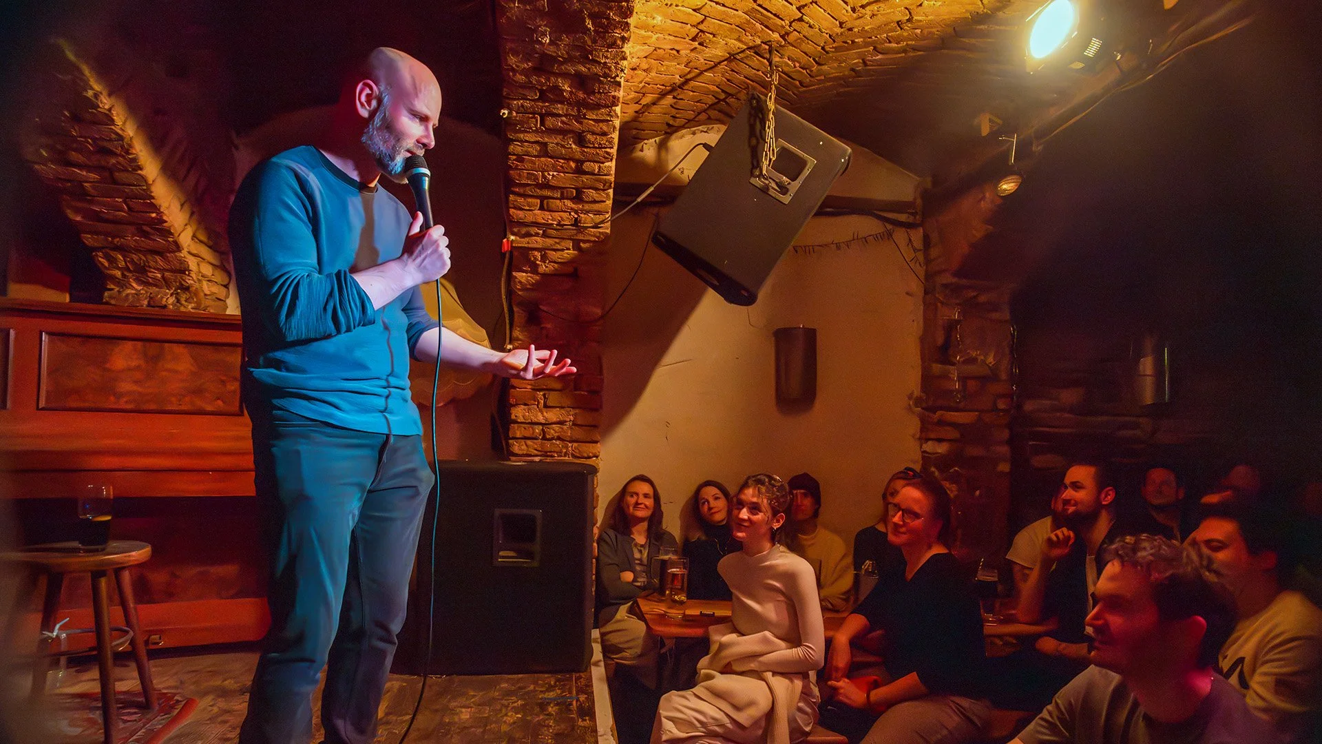 Host Brendan Hickey talking to a smiling front-row audience in a brick-vaulted comedy room in Berlin