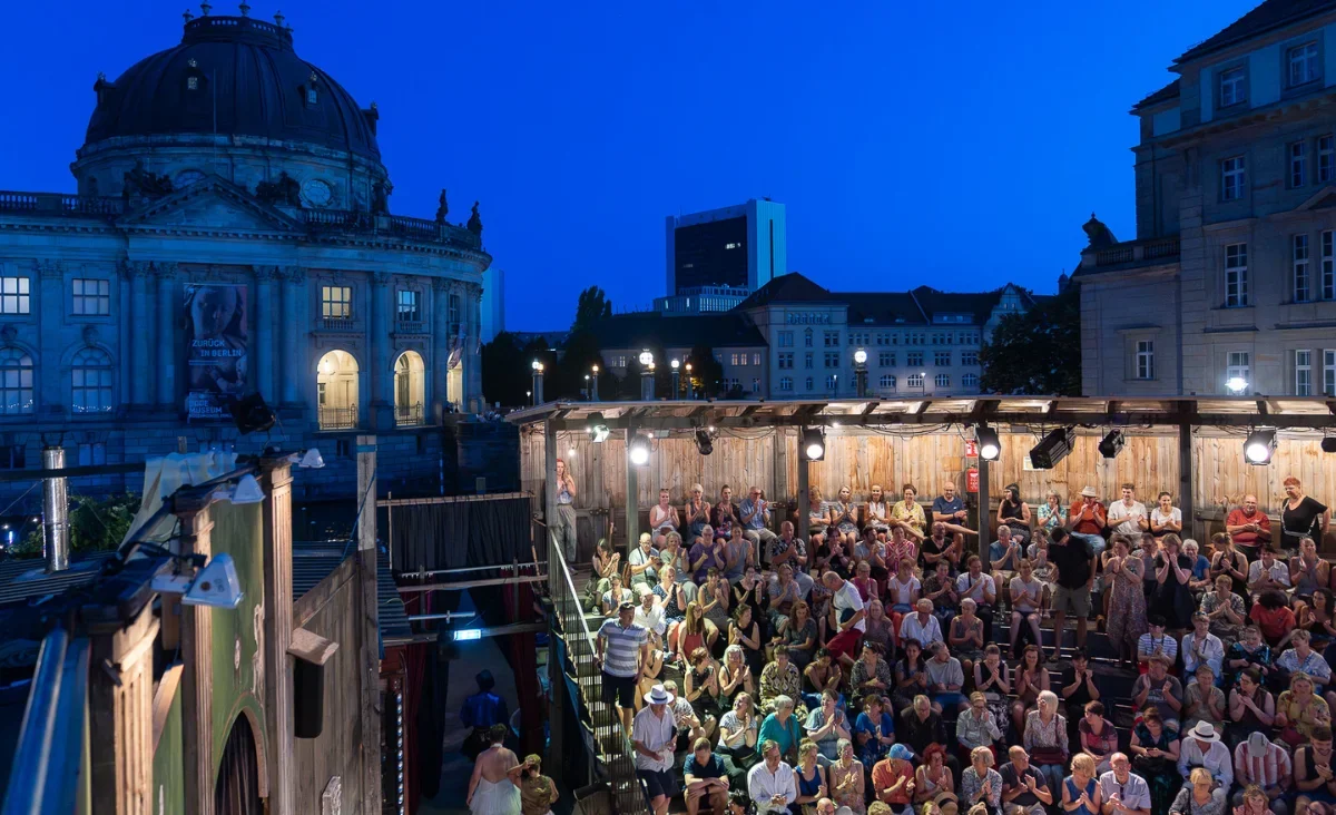 Audience seated at the open-air Monbijou Theater at dusk, with the domed Bode Museum on Museum Island lit up behind, in Berlin