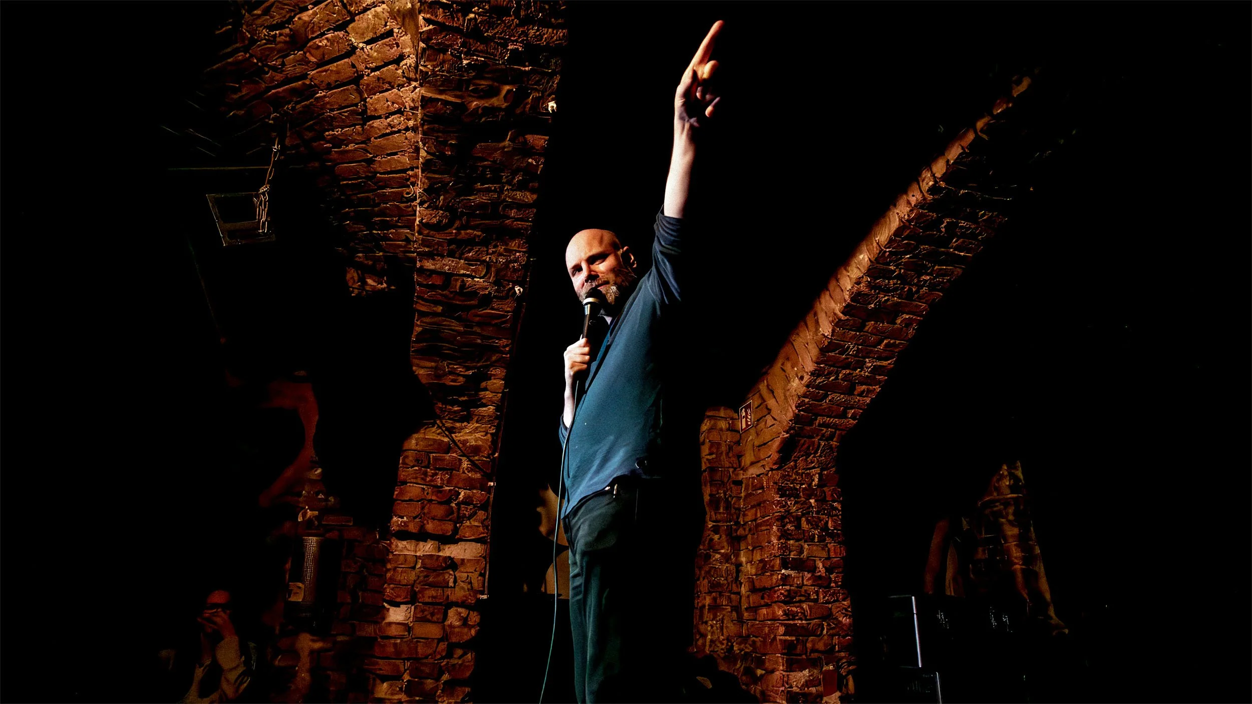 Brendan Hickey (stand-up comedian) on stage at a Berlin comedy show, holding a microphone and pointing upward under a spotlight against an exposed brick wall