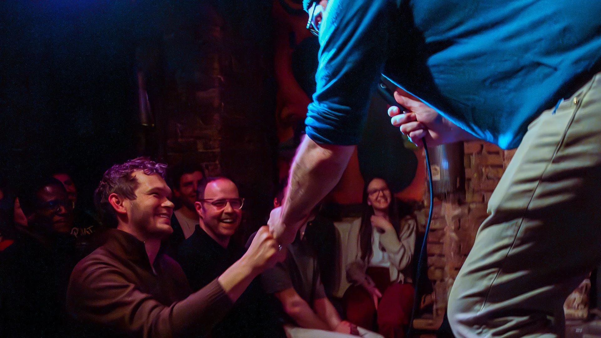 A comedian fist-bumps a laughing front-row audience member during crowd work at an English comedy show in Berlin