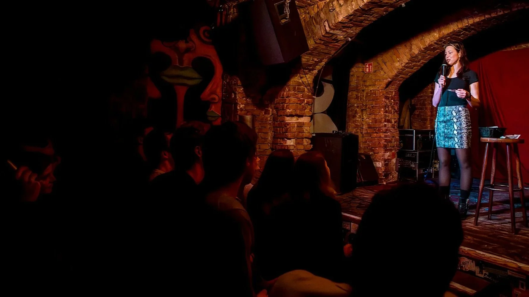 A female comedian reads a note aloud on a dim, brick-arched stage to a silhouetted crowd at a Darkest Thoughts show in Berlin