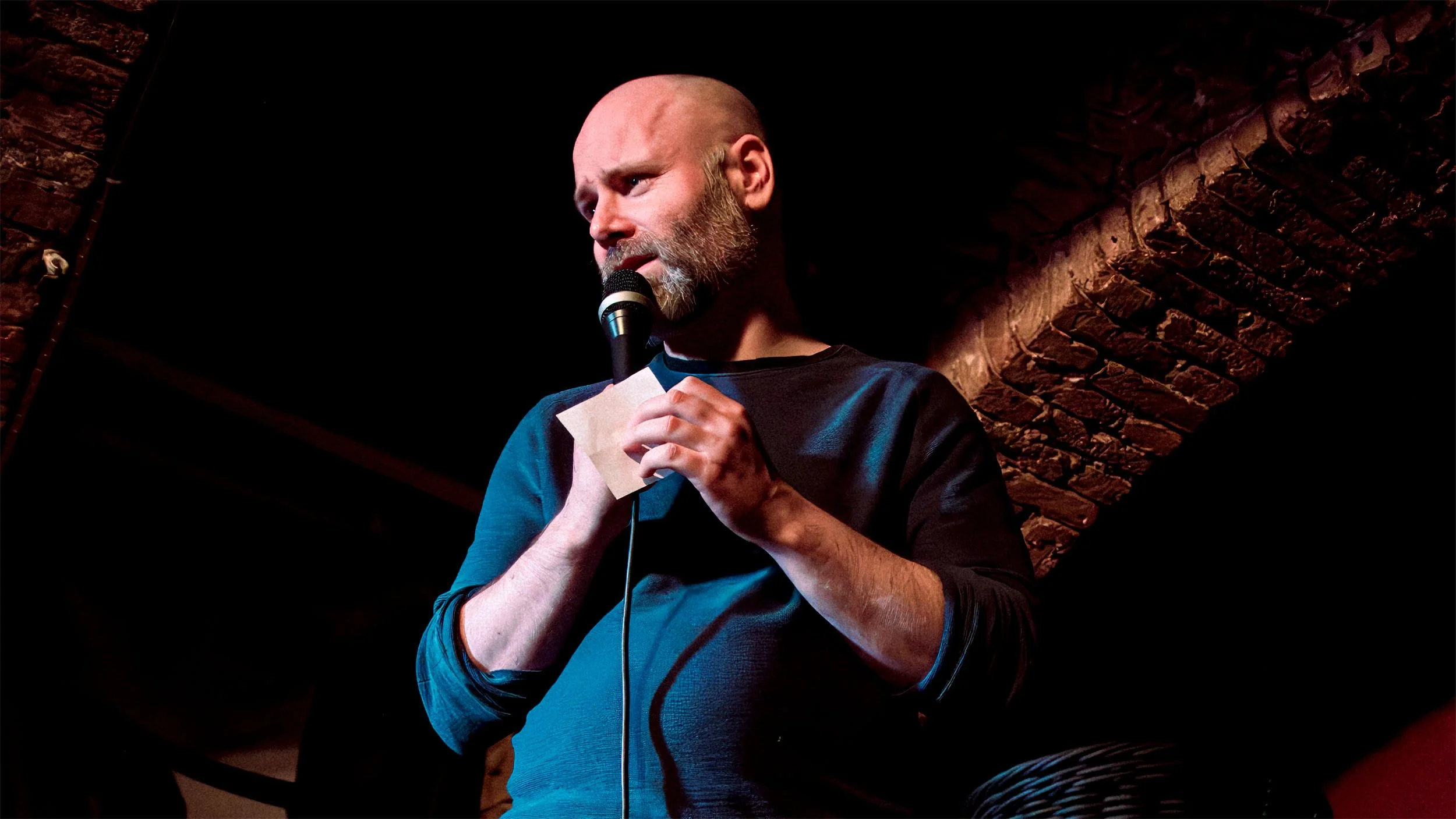 Brendan Hickey (stand-up comedian) reading from a small piece of paper into the microphone on stage at a Berlin comedy night