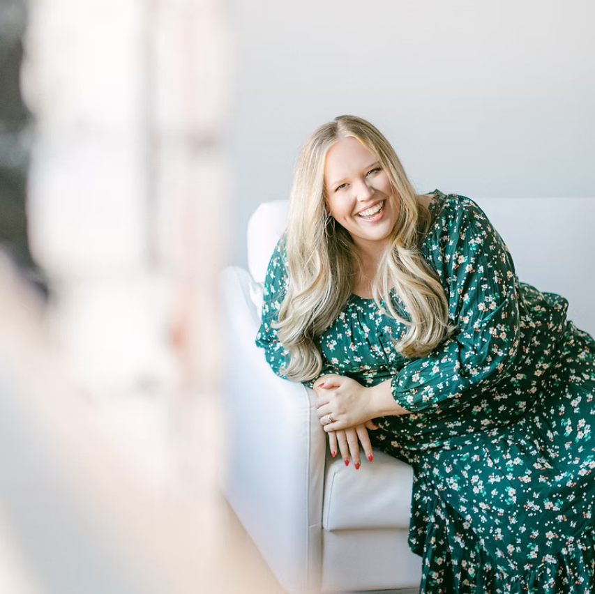 A woman with long blond hair smiling, wearing a teal floral dress and a wide-brimmed beige hat, against a plain brown background.
