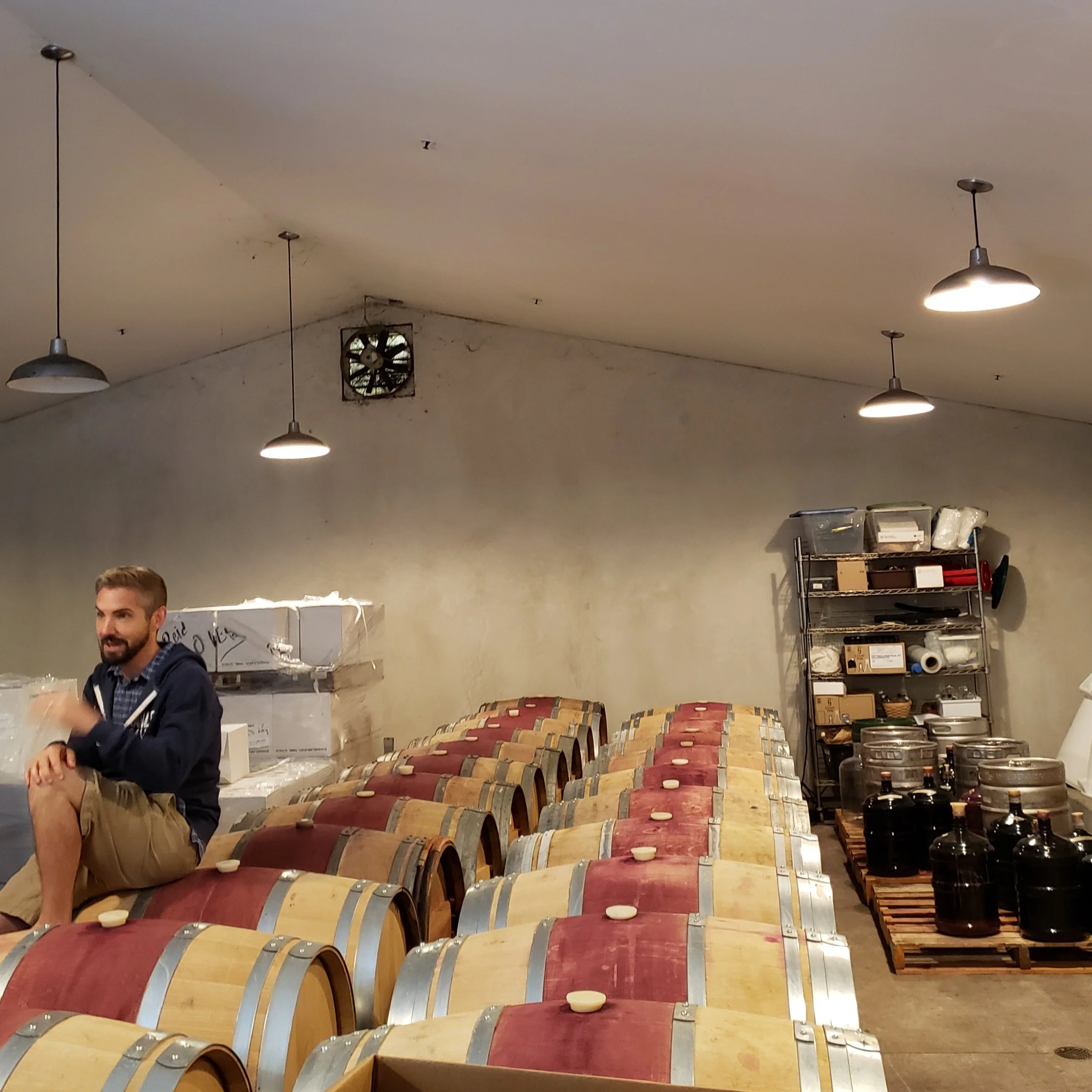 A man sitting on wine barrels in a warehouse or winery storage room, with shelves holding wine bottles and containers behind him.