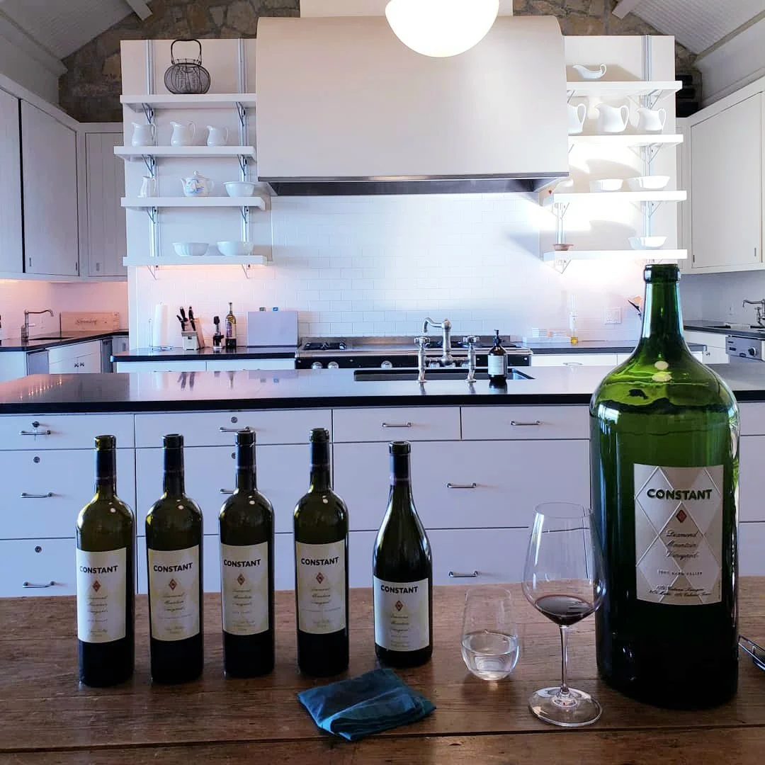 A kitchen with open shelves holding white kitchenware, and a countertop with a wine bottle, wine glass, and five more bottles of wine on a table.