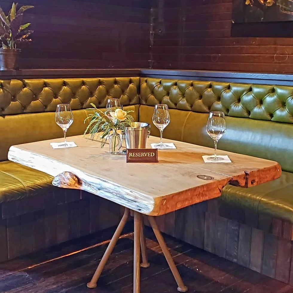 A restaurant table with three empty wine glasses, a flower arrangement, a reserved sign, and a small metal bucket, surrounded by green upholstered seating against wooden walls.