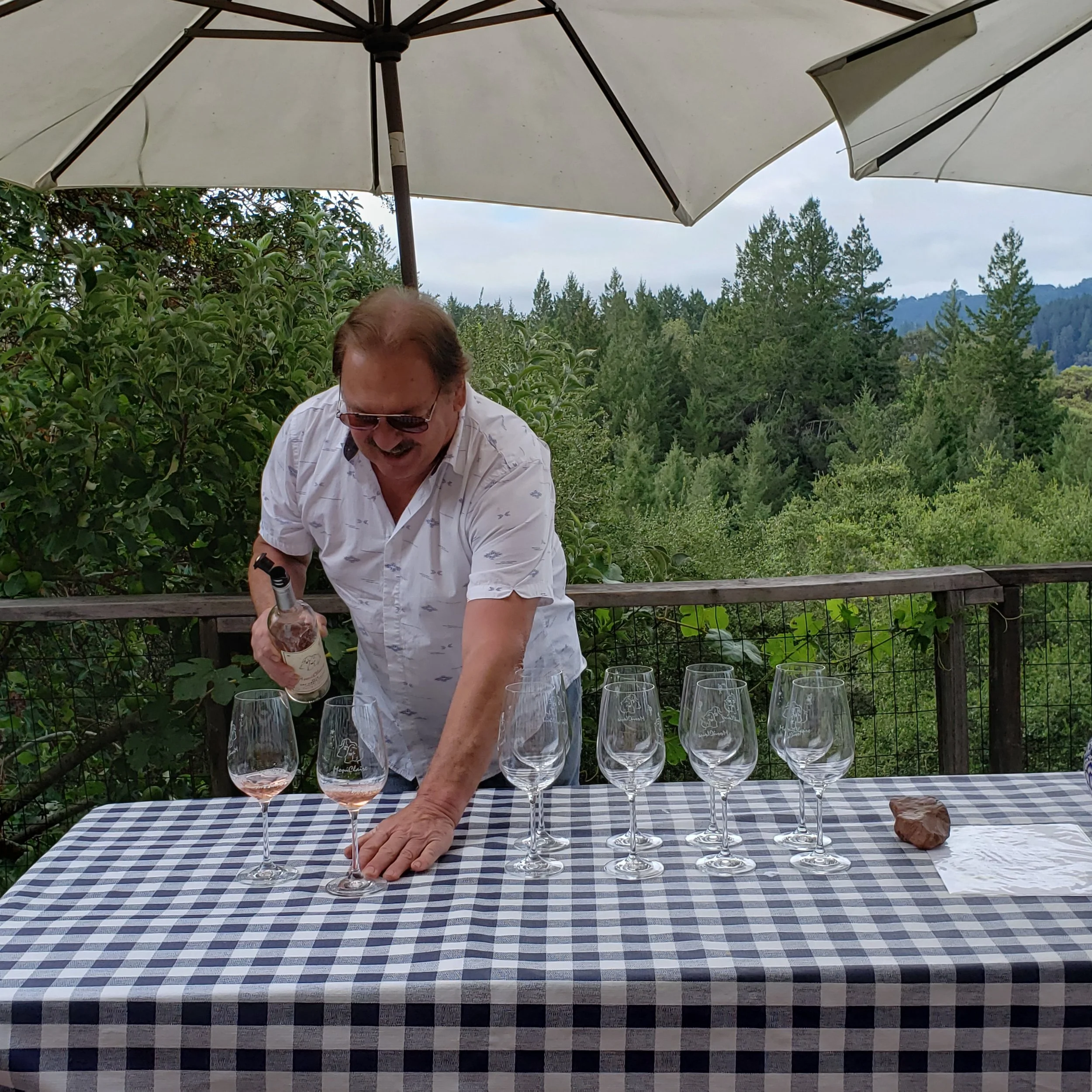 A man with sunglasses and a white short-sleeve shirt standing behind a table with a black and white checkered tablecloth, pouring rosé wine into glasses on an outdoor deck with lush green trees and mountains in the background.