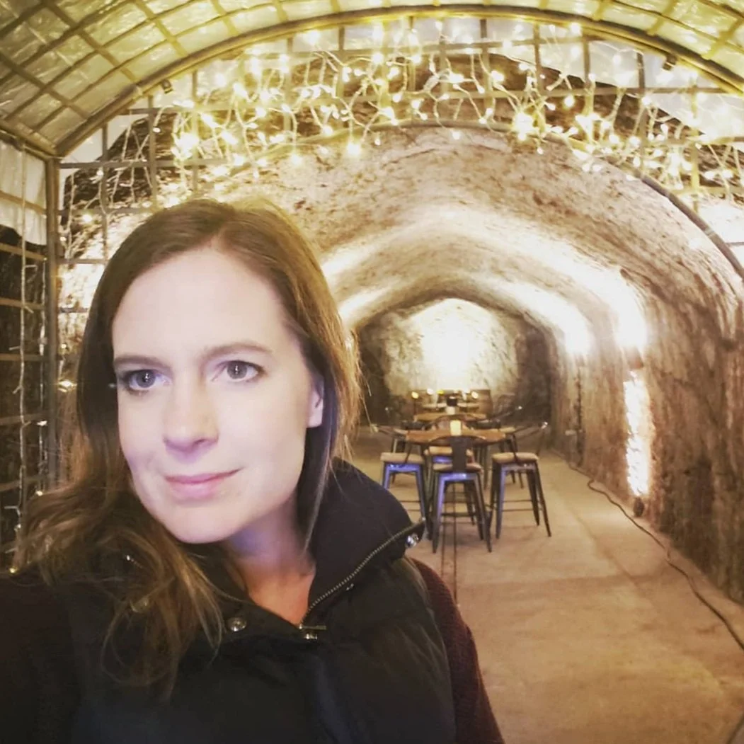 A woman with shoulder-length brown hair taking a selfie inside a tunnel-like cellar with stone walls. Decorative string lights are hanging on the ceiling, illuminating a dining area with tables and chairs in the background.