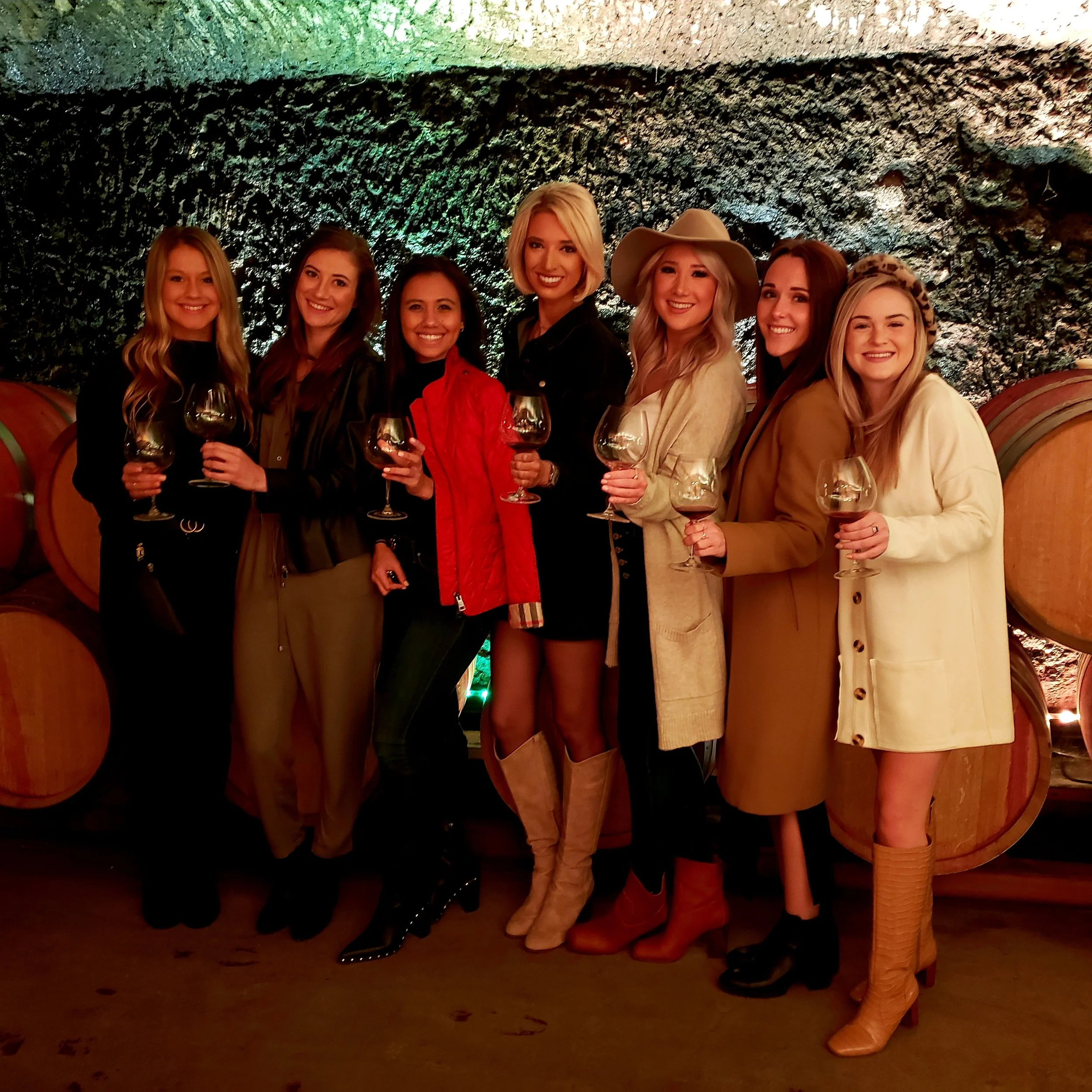 Seven women standing side by side in a wine cellar, holding glasses of red wine and smiling at the camera.