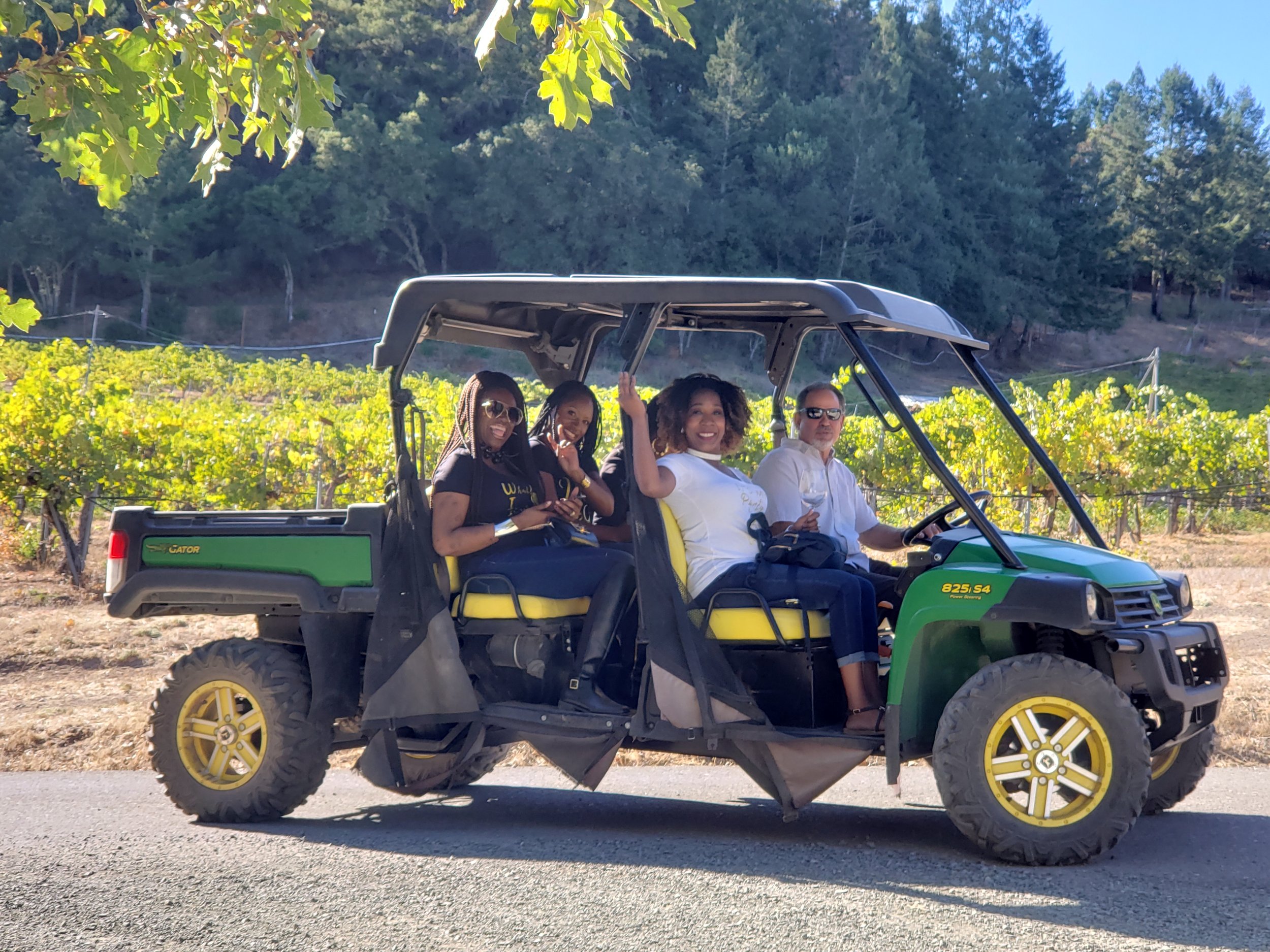 Four people sitting in a green and black golf cart on a vineyard with green vines and trees in the background. The driver is a woman in white, the passenger is a man in sunglasses, and two women are seated behind them, one in black and one in a black