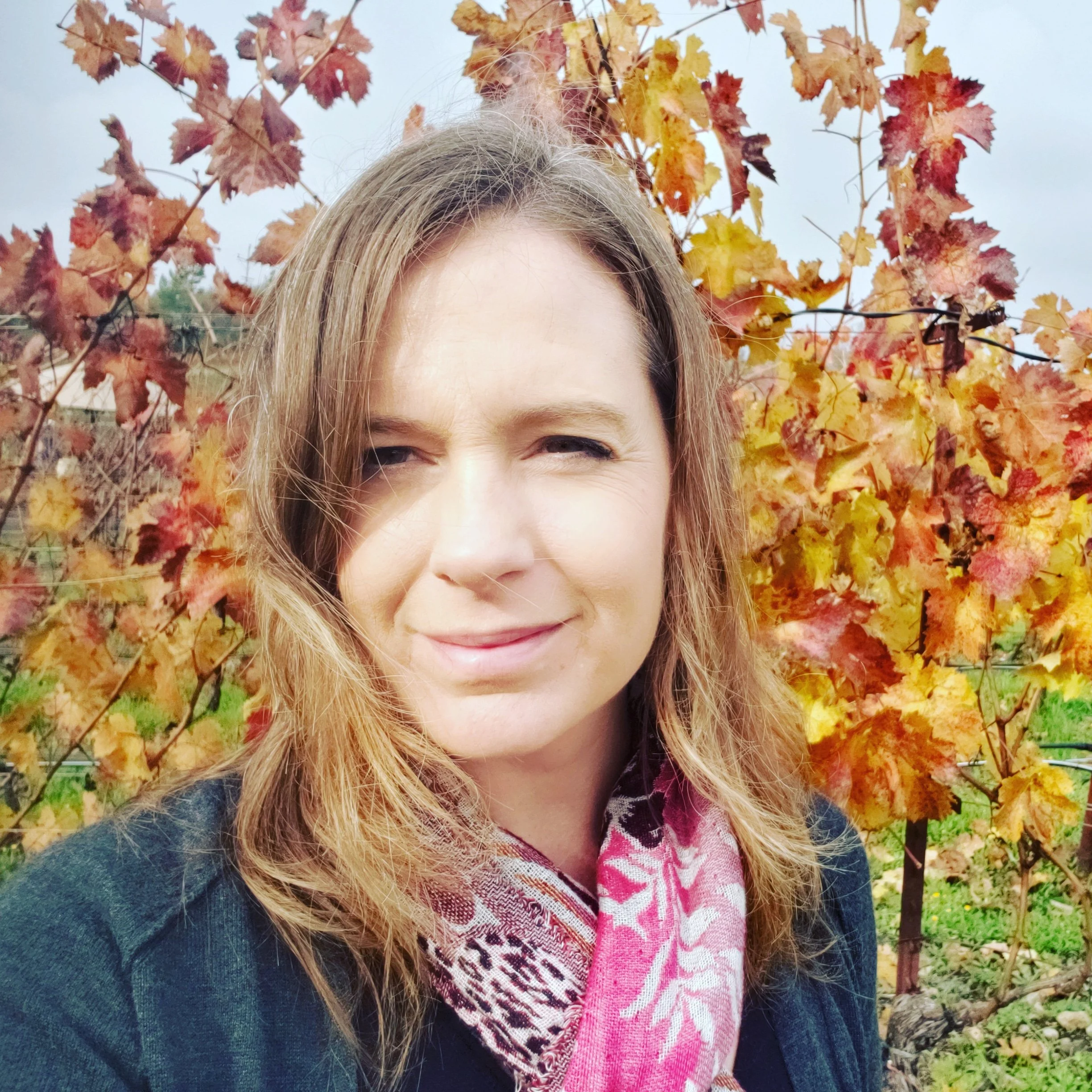 A woman outdoors with a background of autumn-colored leaves, wearing a black top and a pink patterned scarf.