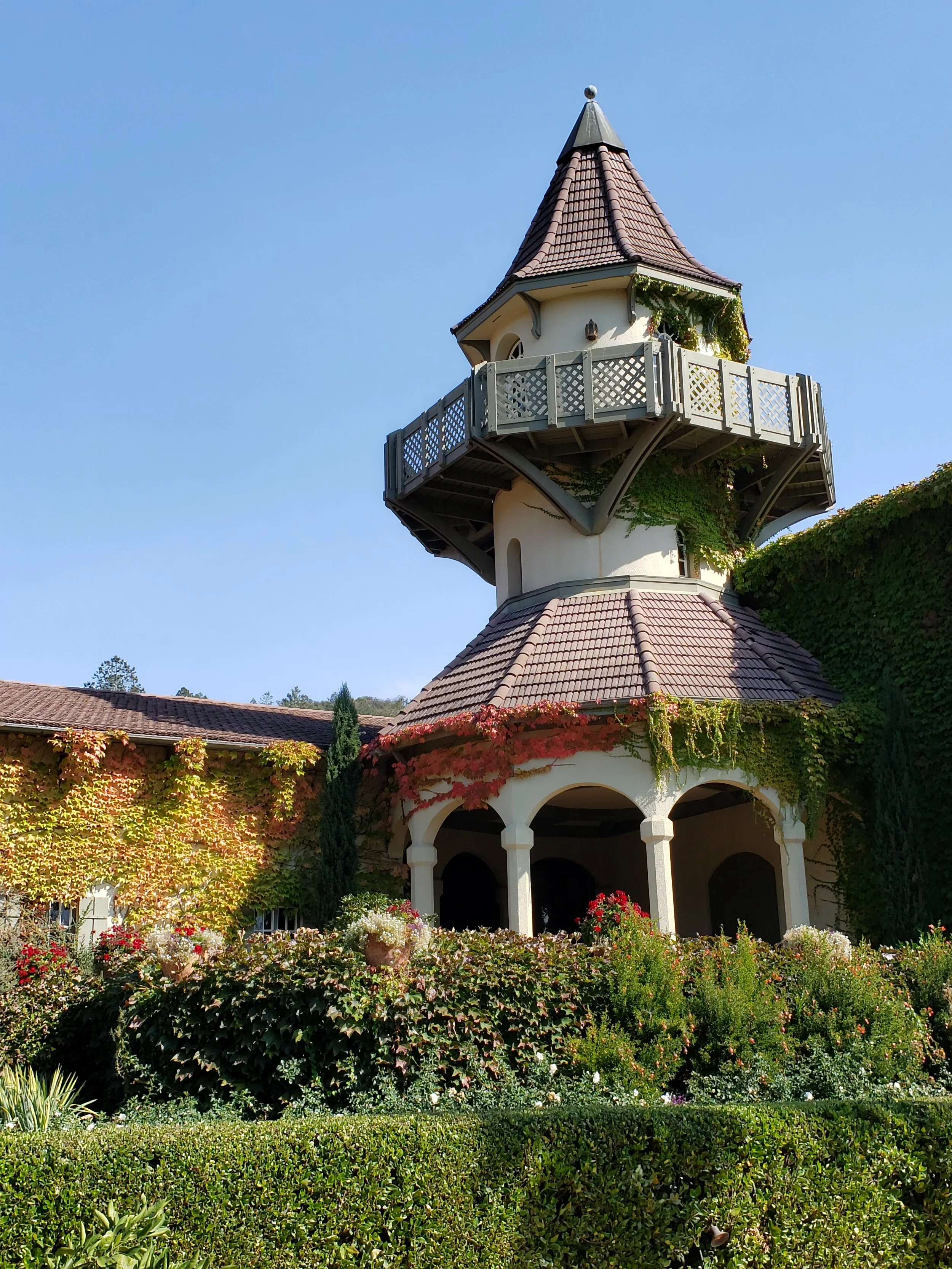 A whimsical multi-story tower with a cone-shaped roof, surrounded by lush green plants and colorful flowers, under a clear blue sky.