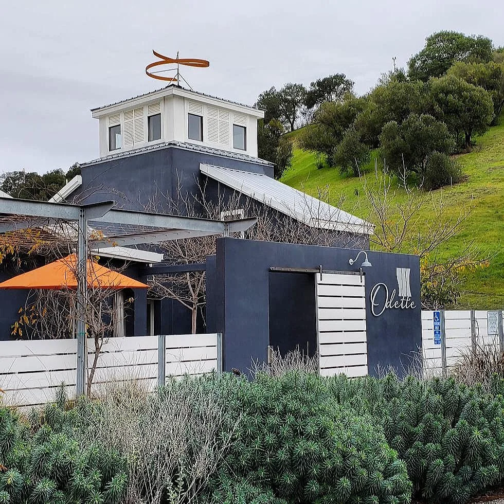 Modern building with a sign that says 'Odette', outdoor seating with an orange umbrella, surrounded by greenery and a hill in the background.