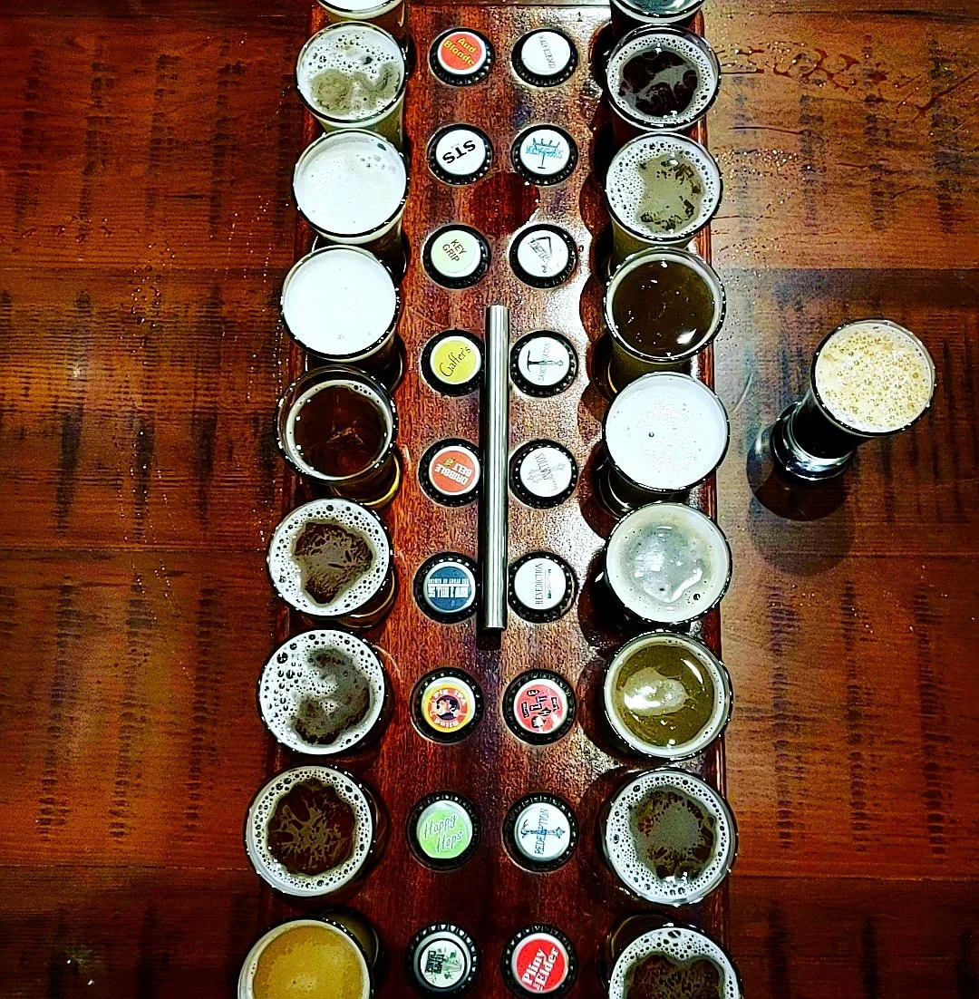 A top-down view of 20 small glasses of various beers arranged on a wooden table with beer bottle caps placed between and around the glasses.
