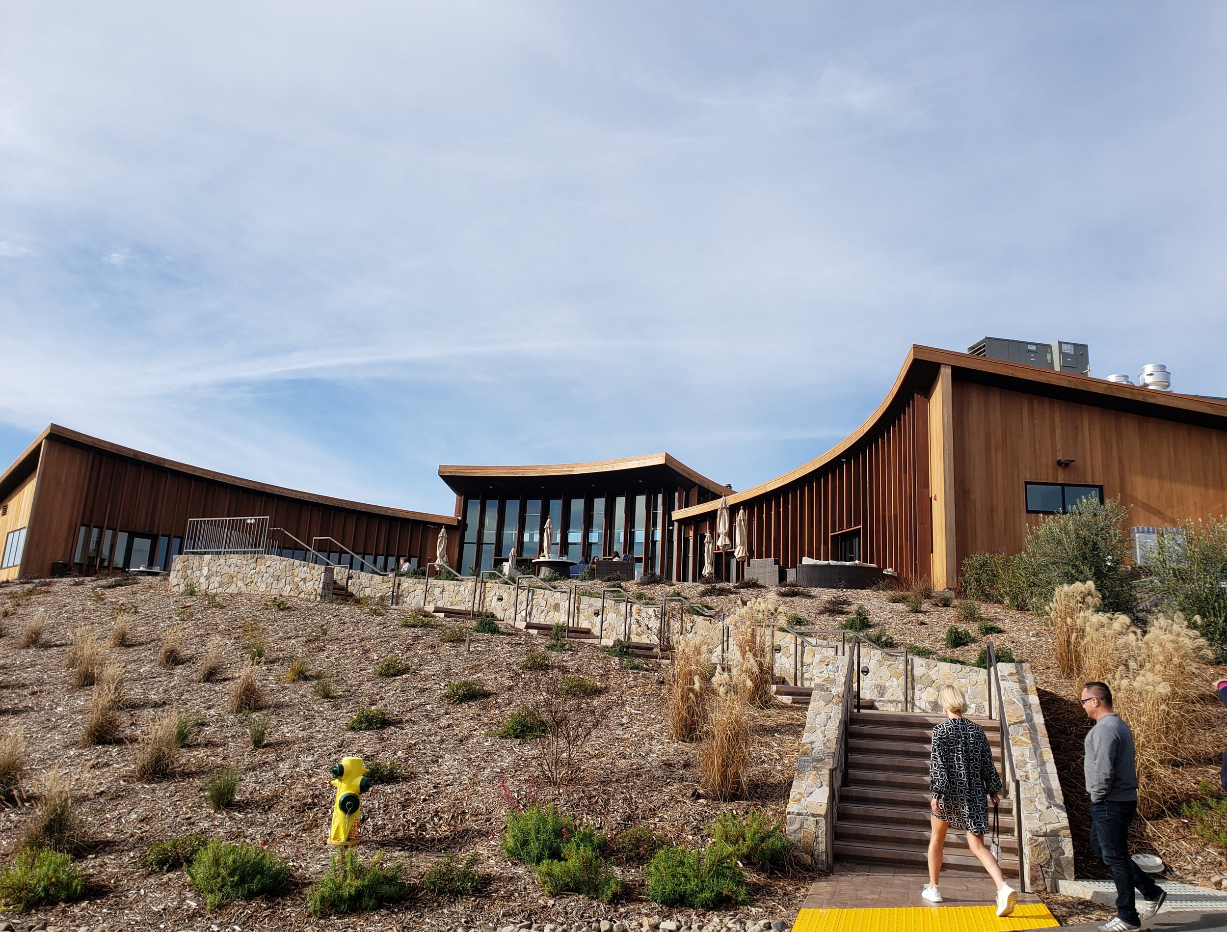 People walking up stairs outside a modern building with a wooden exterior and large glass windows, on a hillside with sparse vegetation, under a partly cloudy sky.