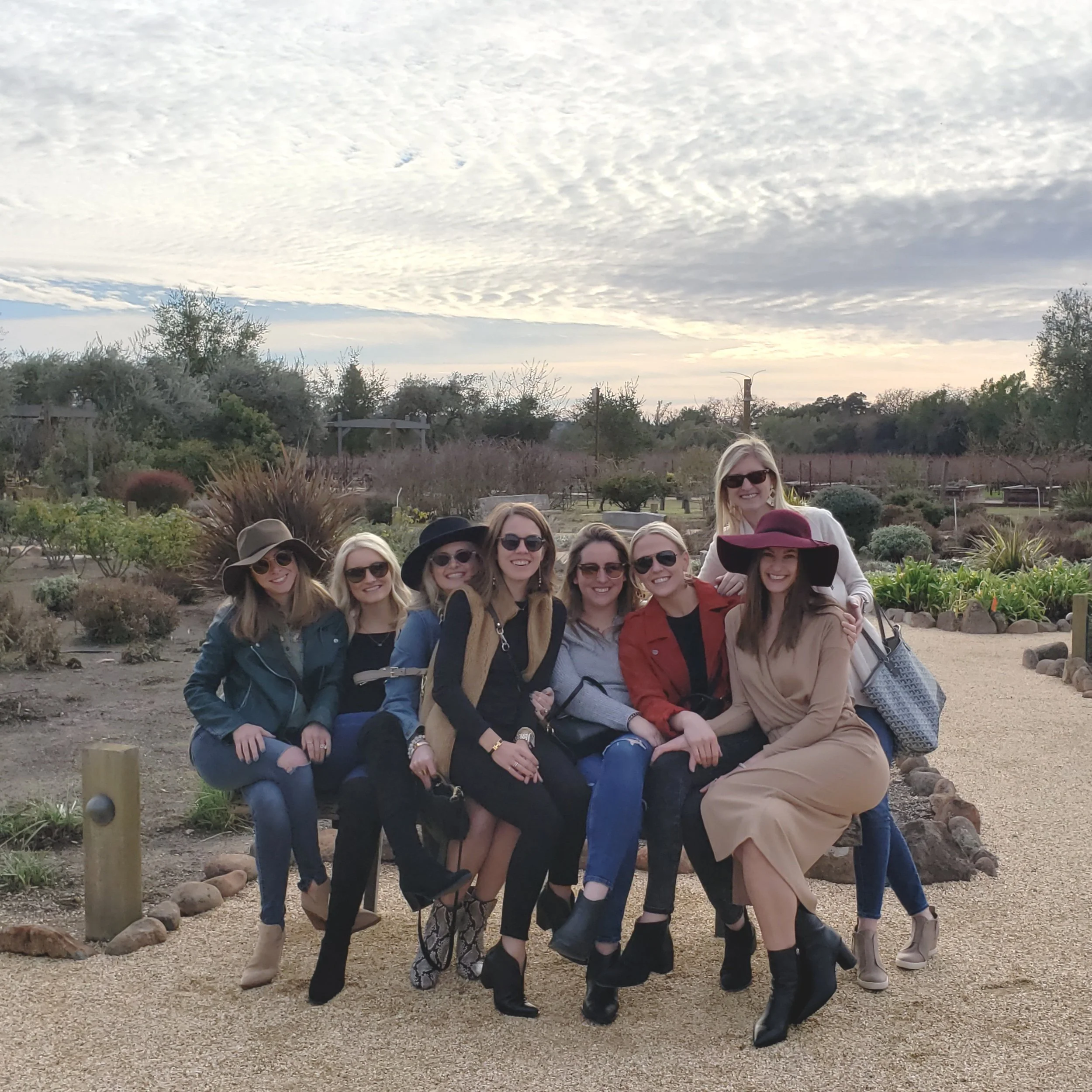 Group of nine women sitting and standing on a bench in a garden, wearing sunglasses and stylish clothing, smiling at the camera during daytime.