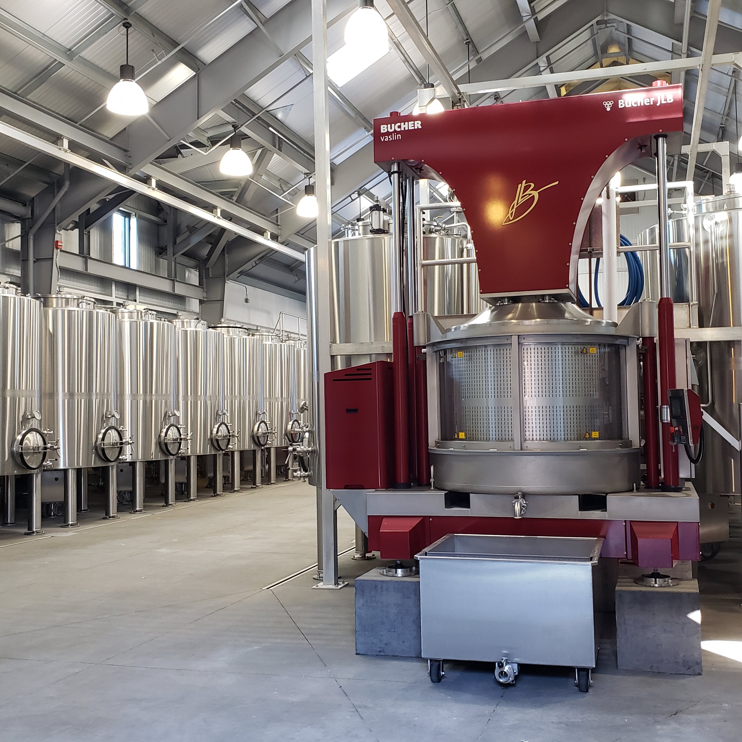 Inside a winery, large stainless steel tanks for wine fermentation and a red bottling machine in the foreground.