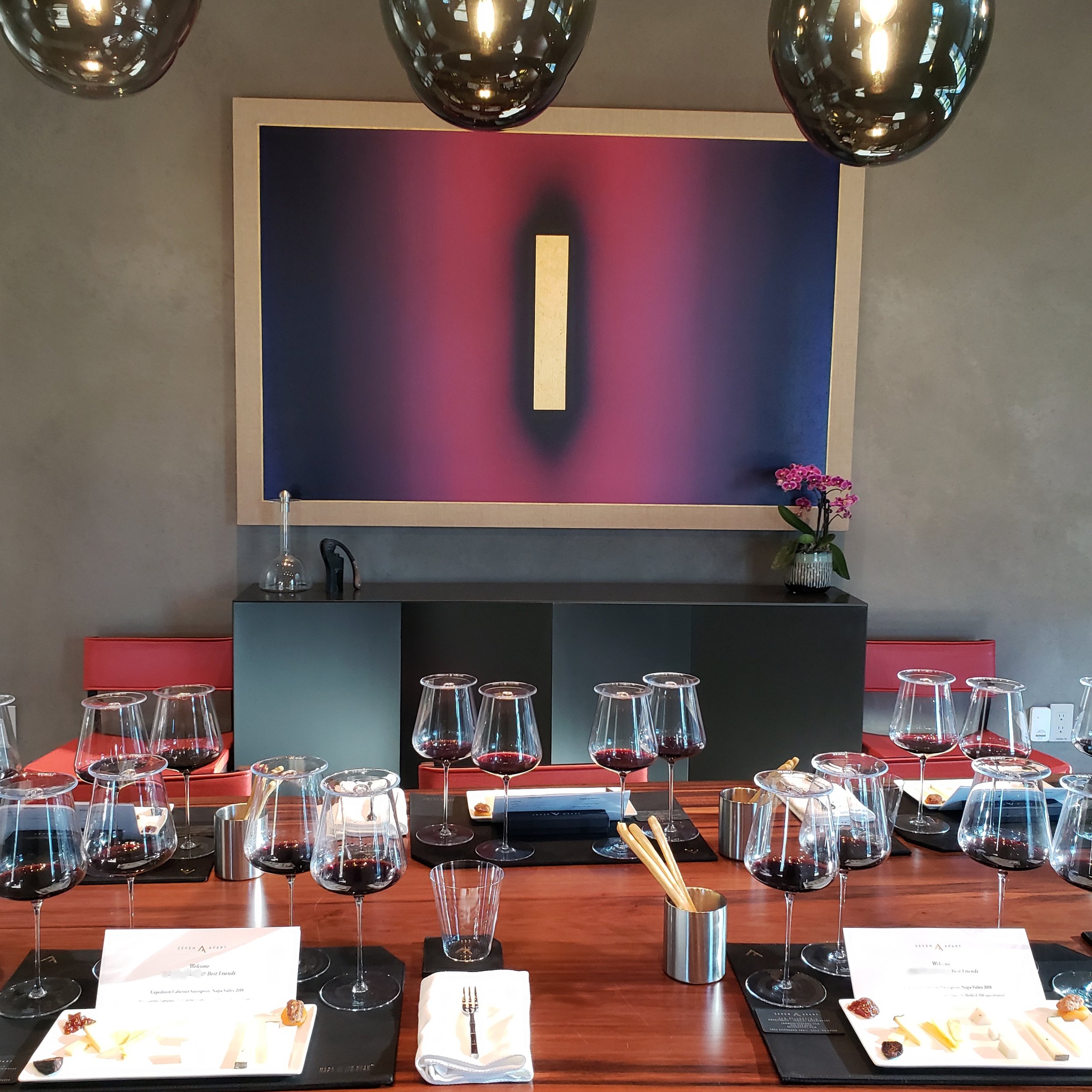 A table set for a wine tasting with multiple glasses of red wine, tasting notes, and tasting tools, in a modern room with abstract artwork on the wall.