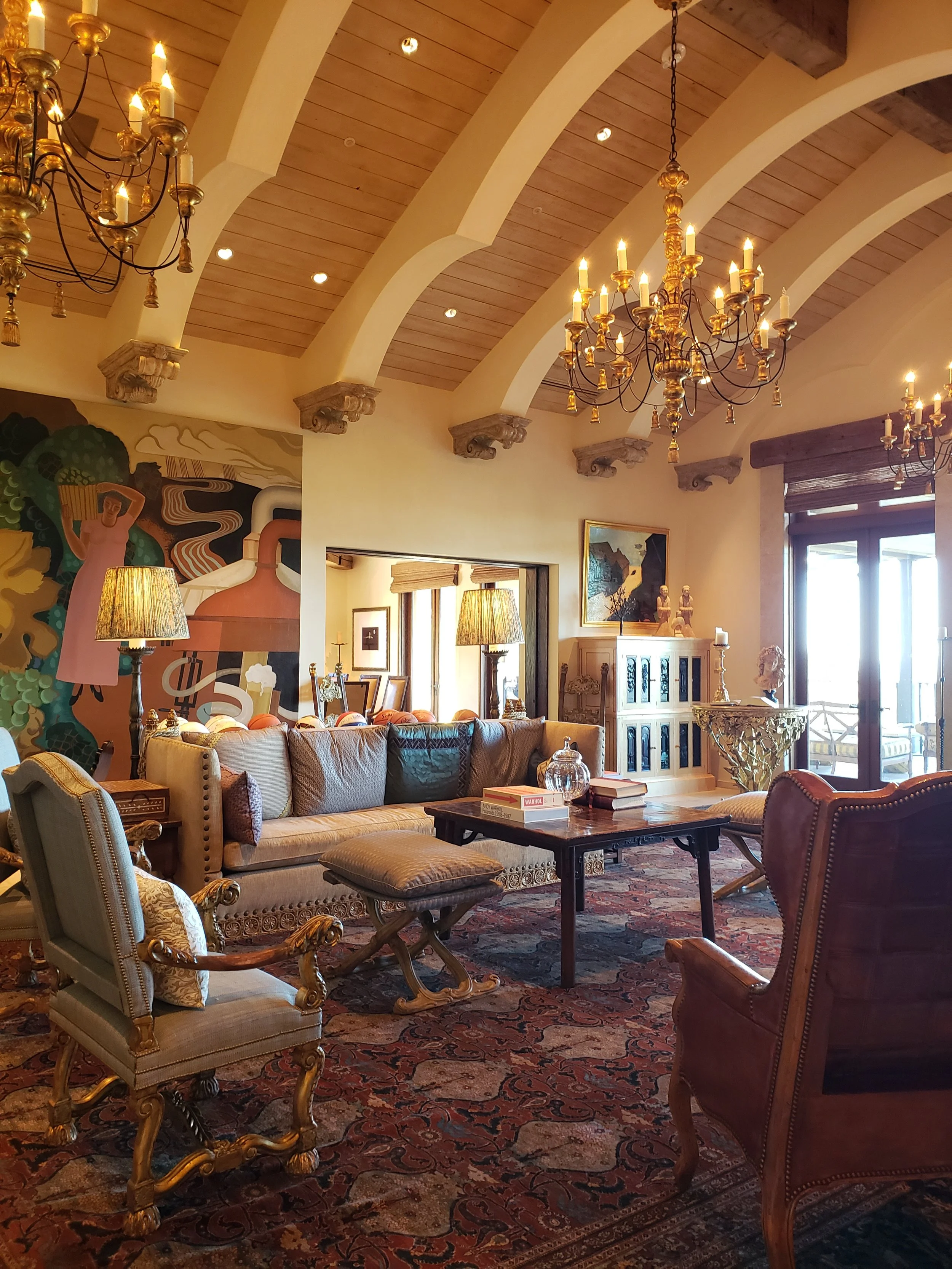 Elegant living room with ornate furniture, colorful rug, chandeliers, and artwork, with large windows and natural light.