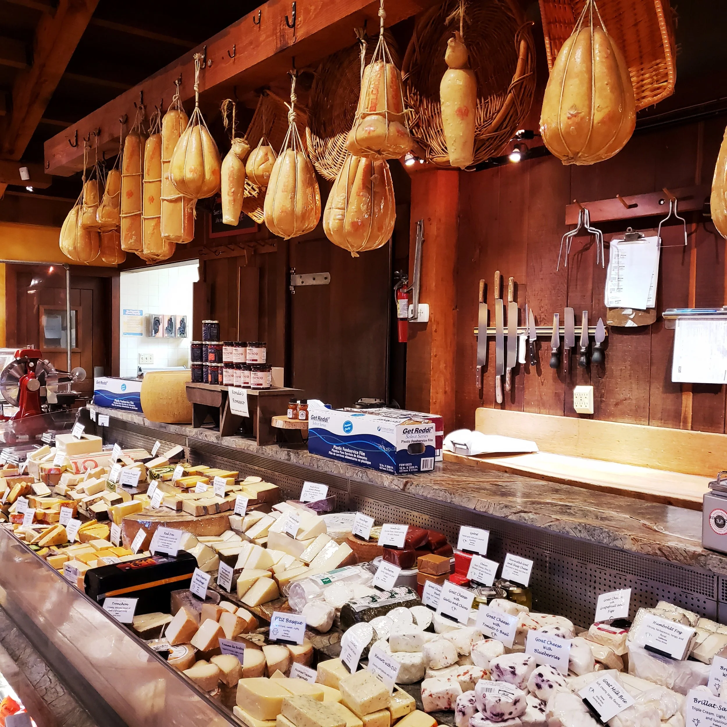 Display of various cheeses on a counter with hanging cured meats above in a rustic cheese shop.